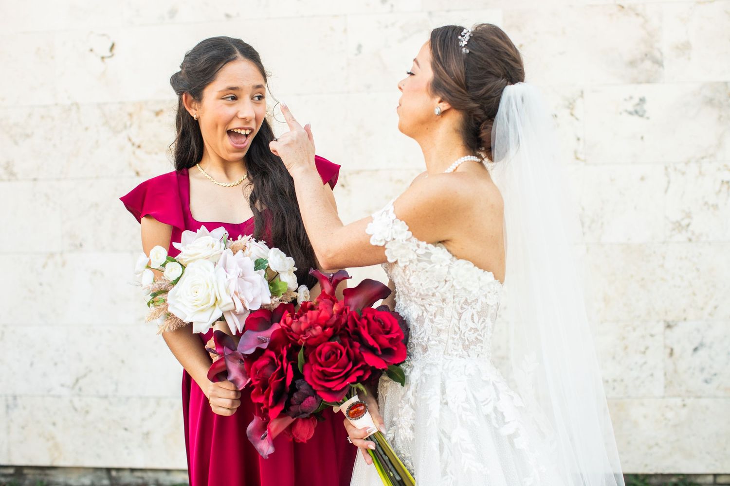 Bride in lace gown laughing with bridesmaid in red dress, both holding bouquets of red and white roses.