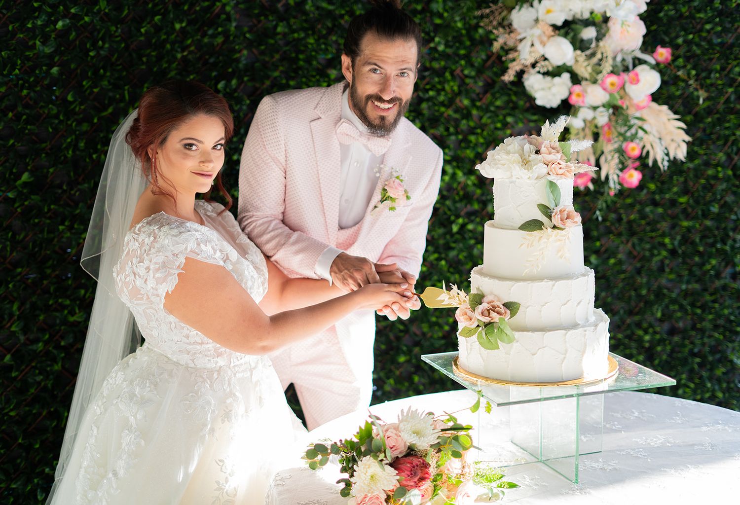 Bride and groom smiling while cutting a elegant four-tier wedding cake adorned with pink flowers outdoors.