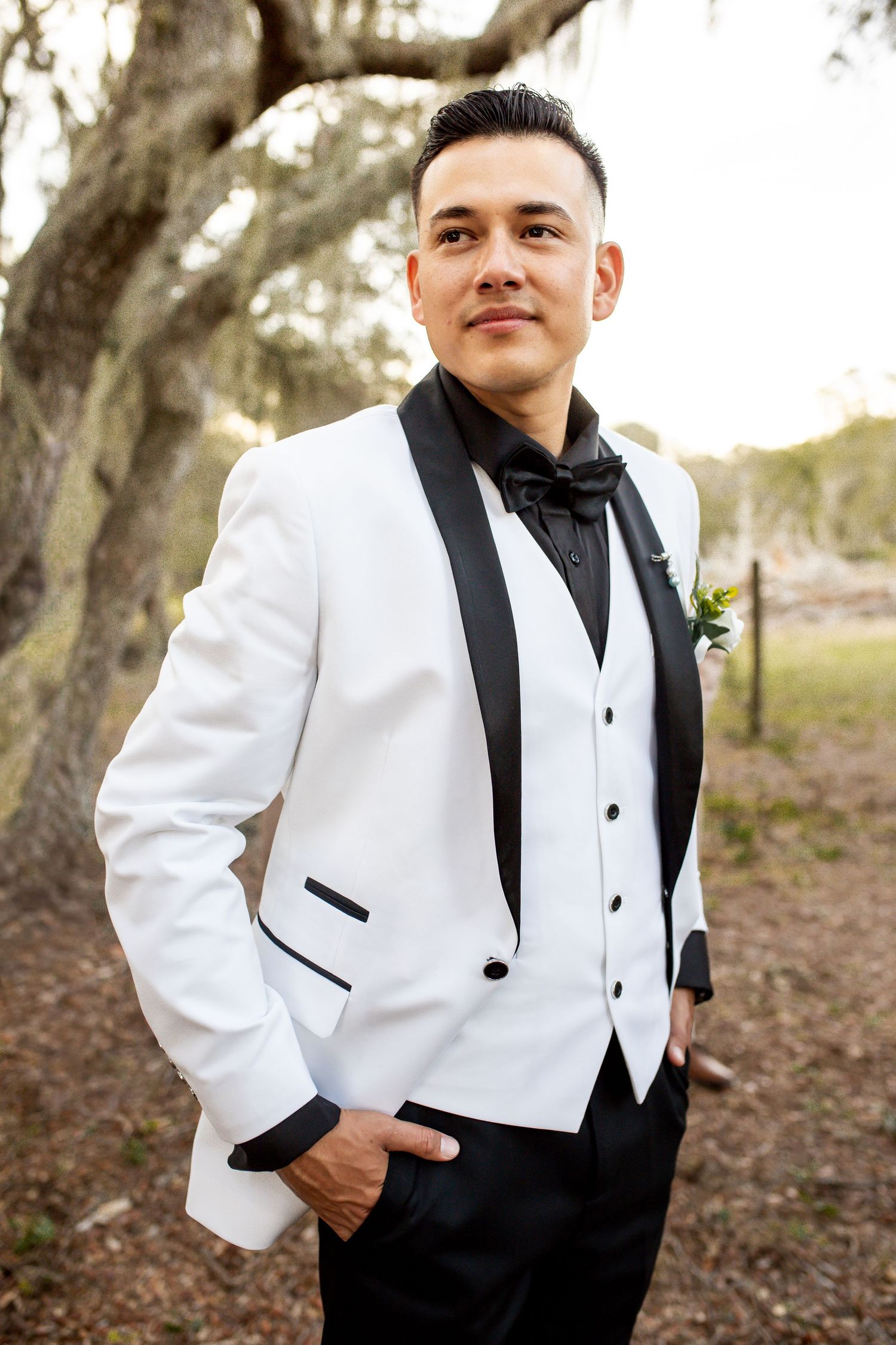 A groom in a classic white tuxedo with black lapels poses against a natural backdrop.
