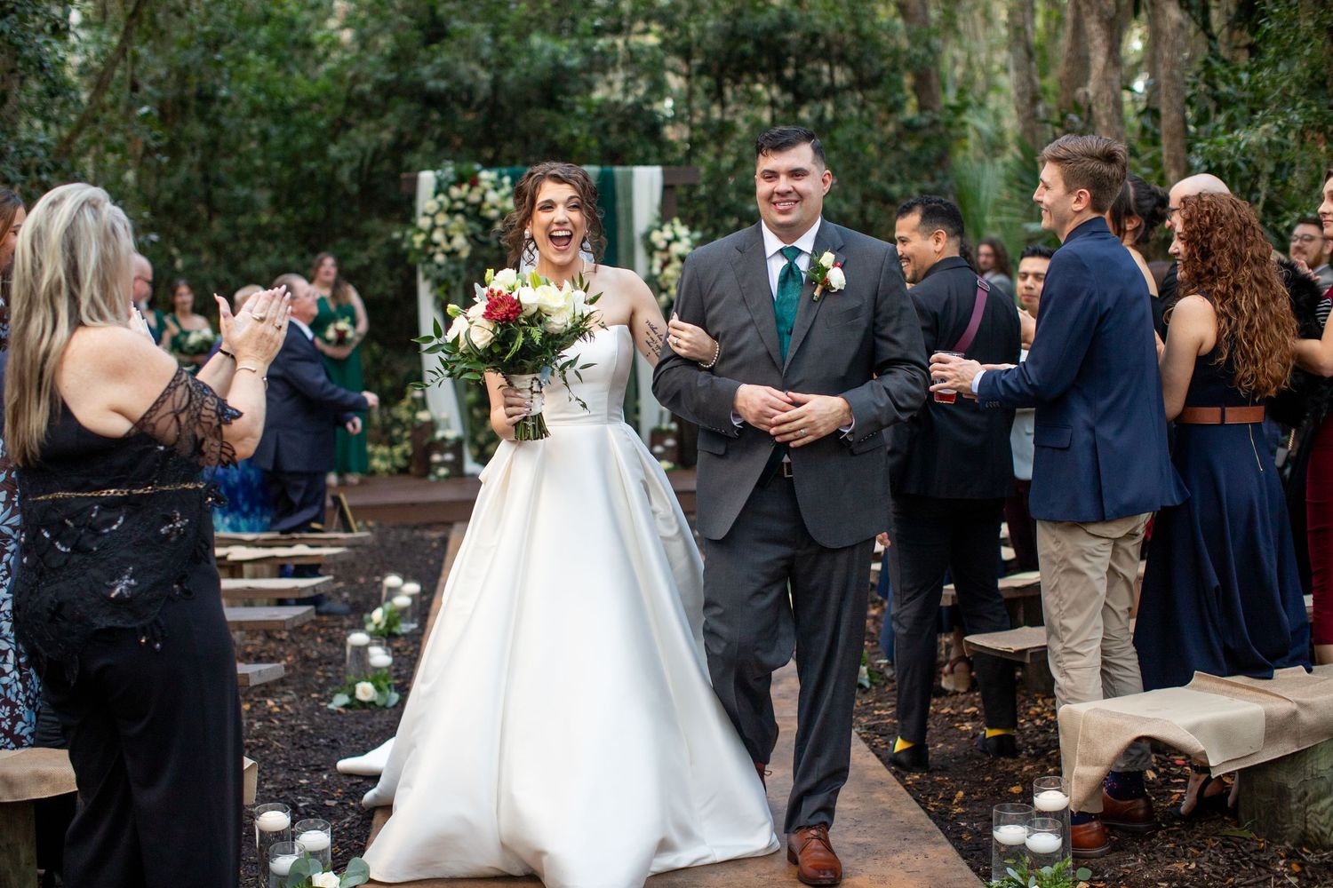 Outdoor garden wedding ceremony with guests seated on wooden benches surrounded by lush greenery.