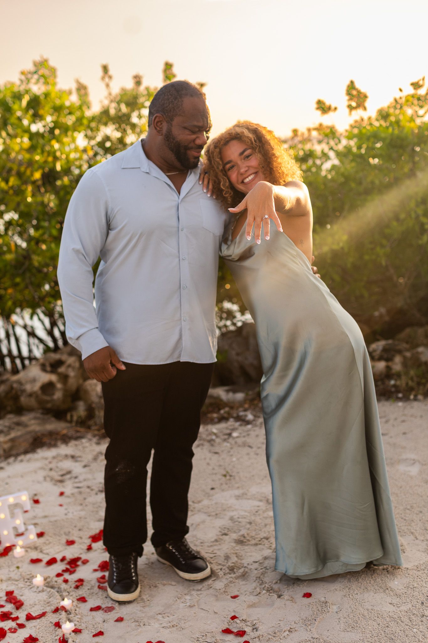 Happy couple celebrating engagement on a sandy beach at sunset, woman showing ring while leaning on man amid rose petals.