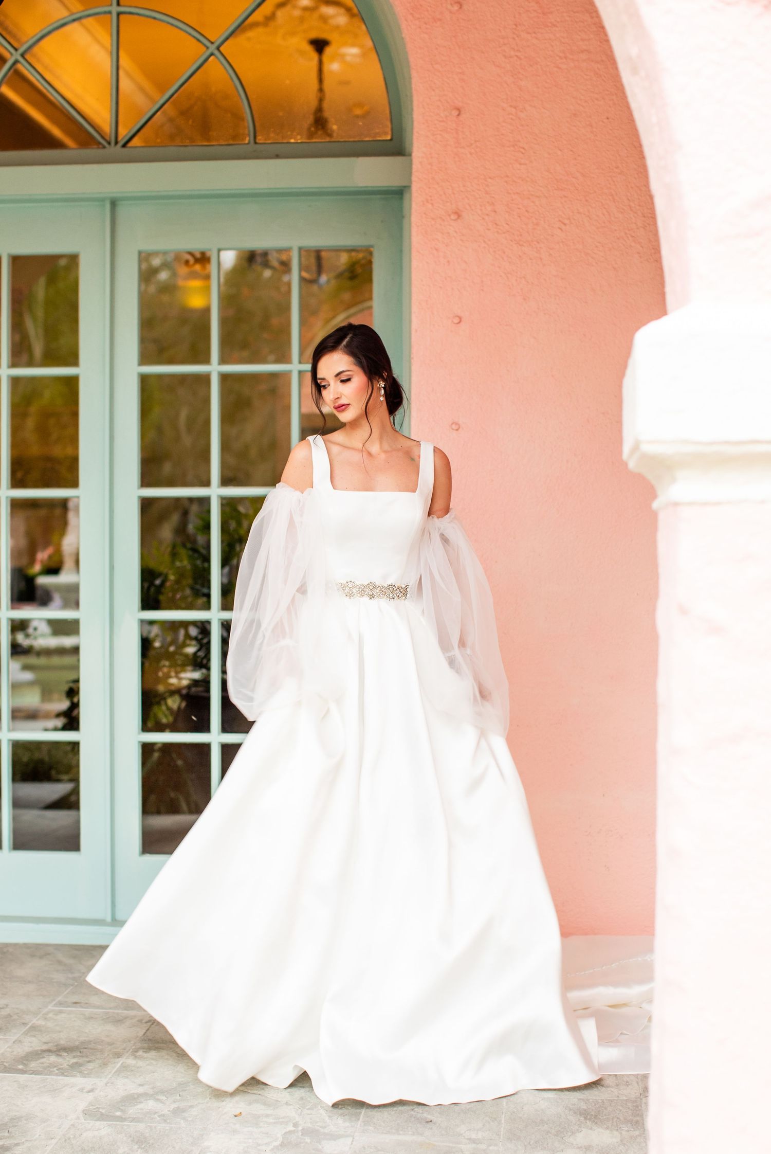 Bride in white ball gown with tulle sleeves and jeweled belt poses by pink stucco arch and mint French doors.