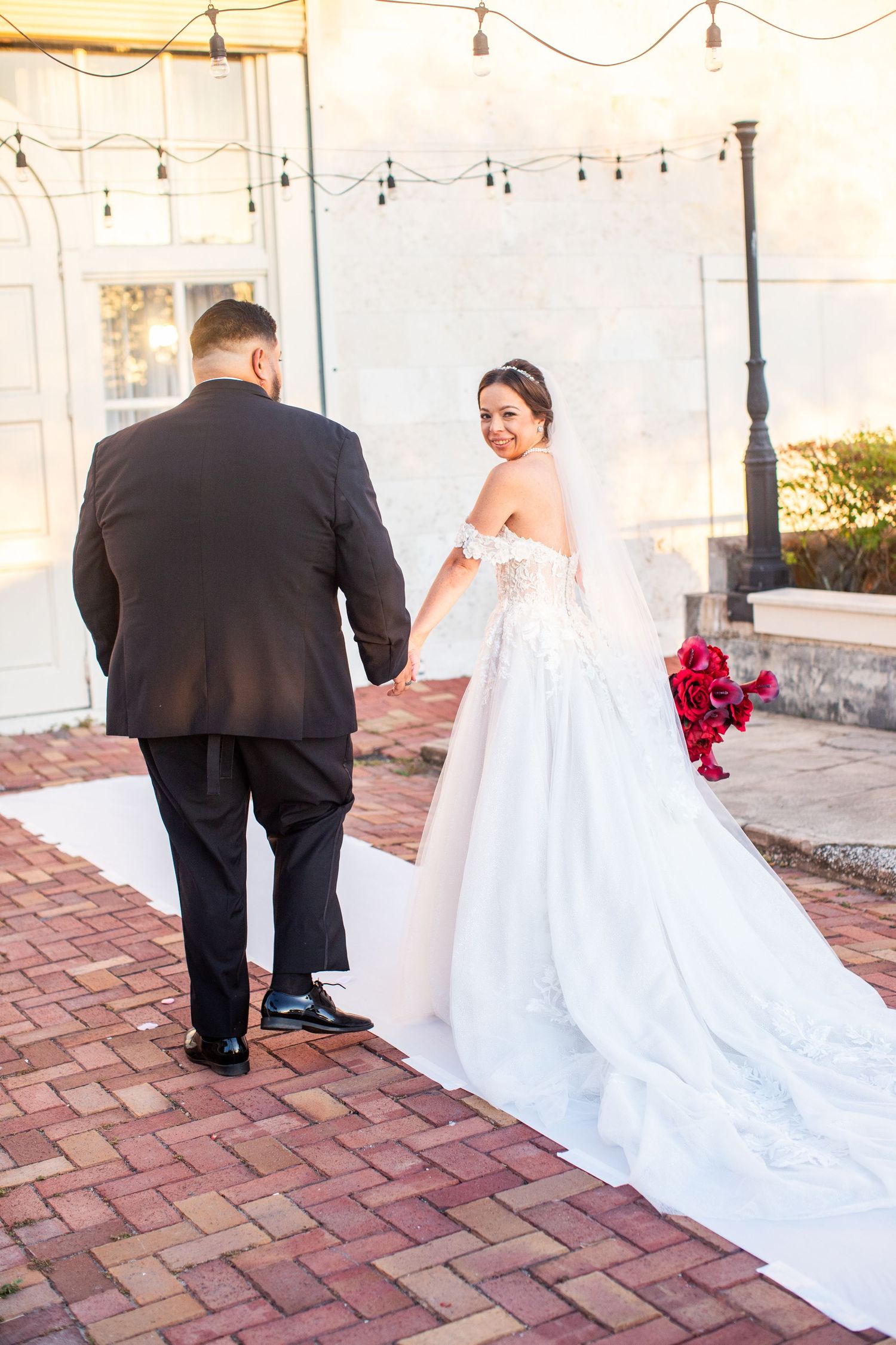 Bride in white off-shoulder gown holding red bouquet walks hand-in-hand with groom in black suit on brick path.