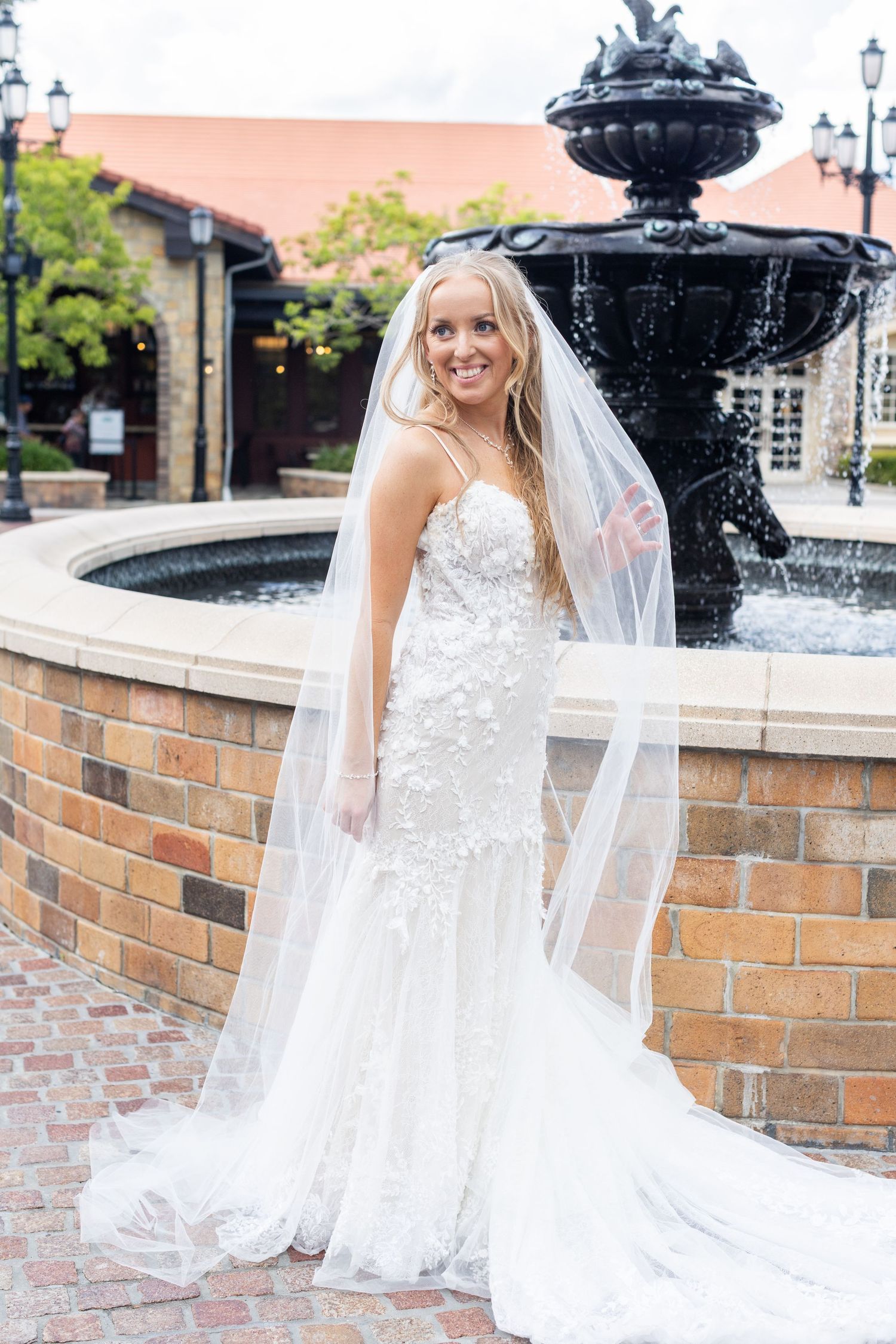 Bride in lace dress poses by brick fountain wall with cascading veil.