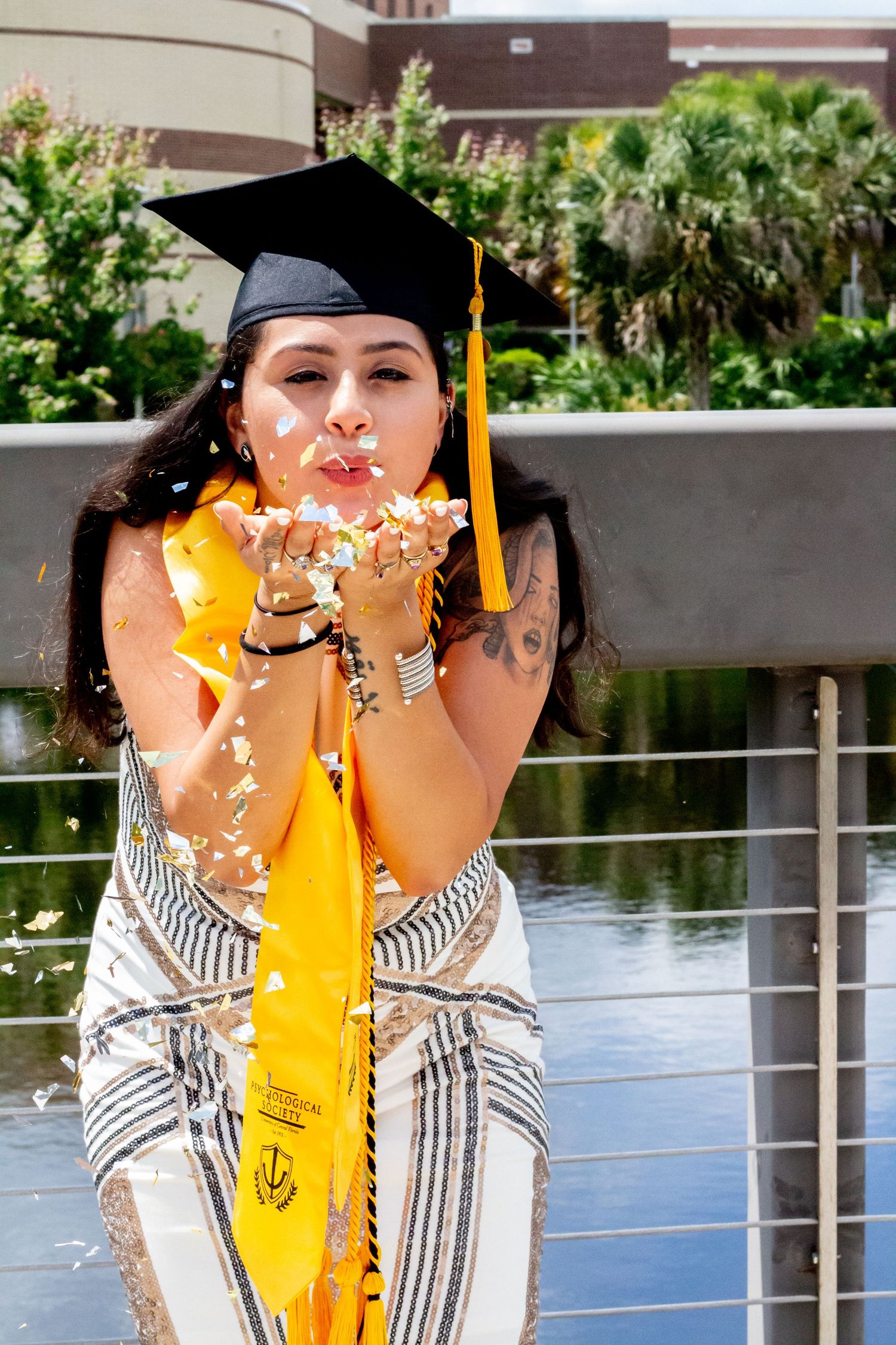 Graduate in cap and yellow stole blows glitter during outdoor photo session.