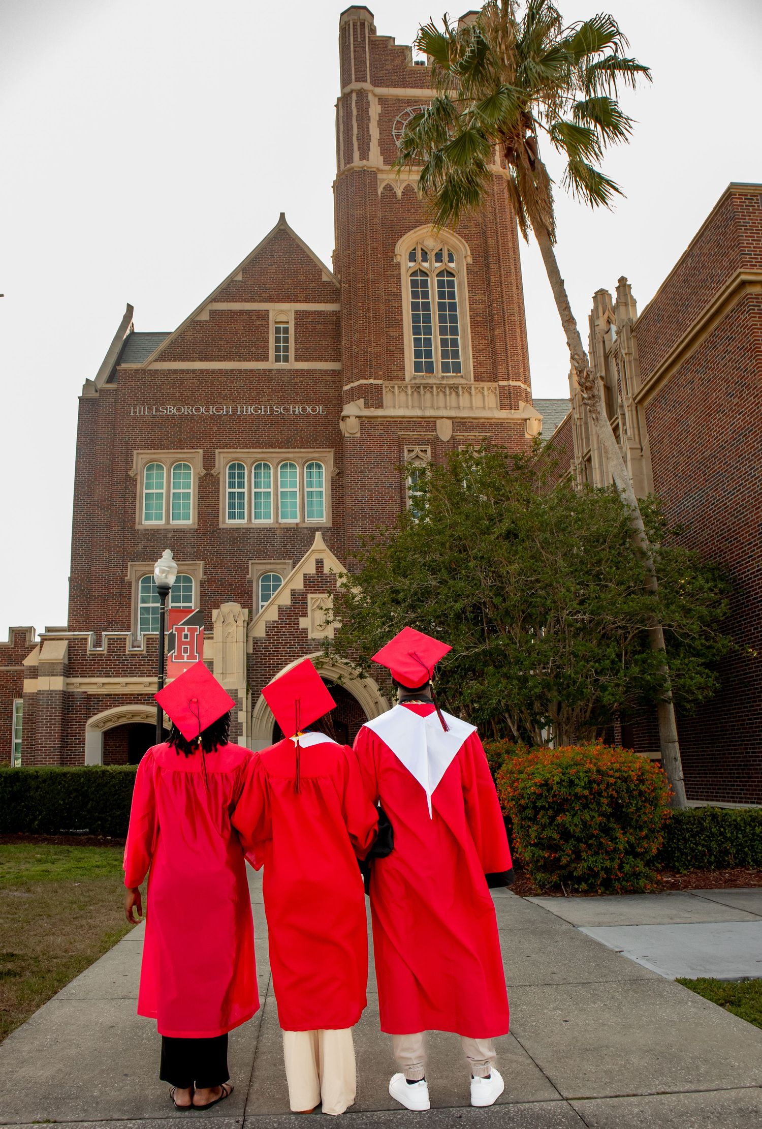 Group in red graduation gowns poses playfully in front of brick church building with palm trees.