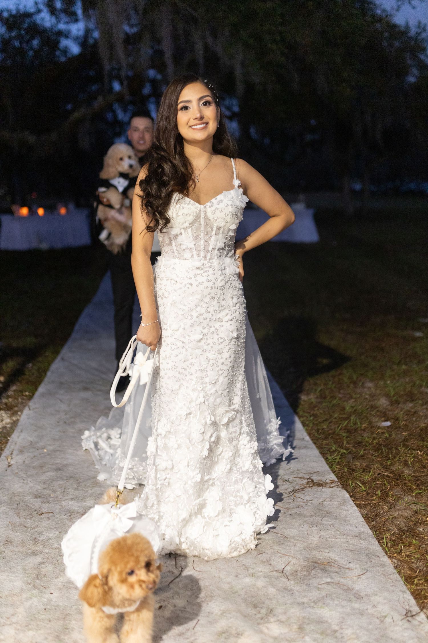 A bride in a white lace gown stands with her small dog on an outdoor path at dusk.