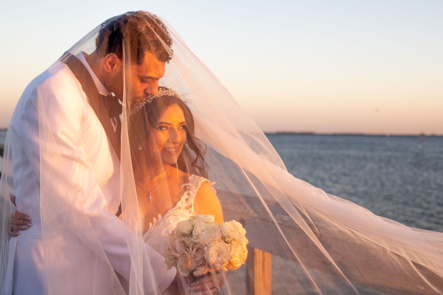 Romantic sunset beach wedding photoshoot with couple embracing as veil flows in the ocean breeze.