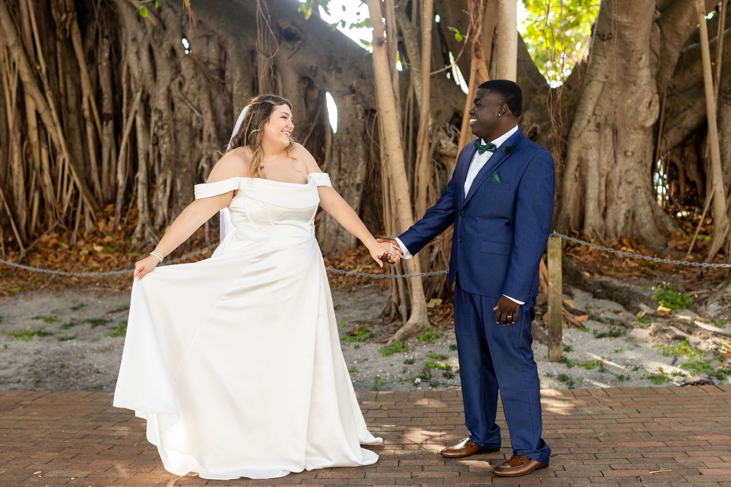 A couple in wedding attire poses playfully under large banyan trees.