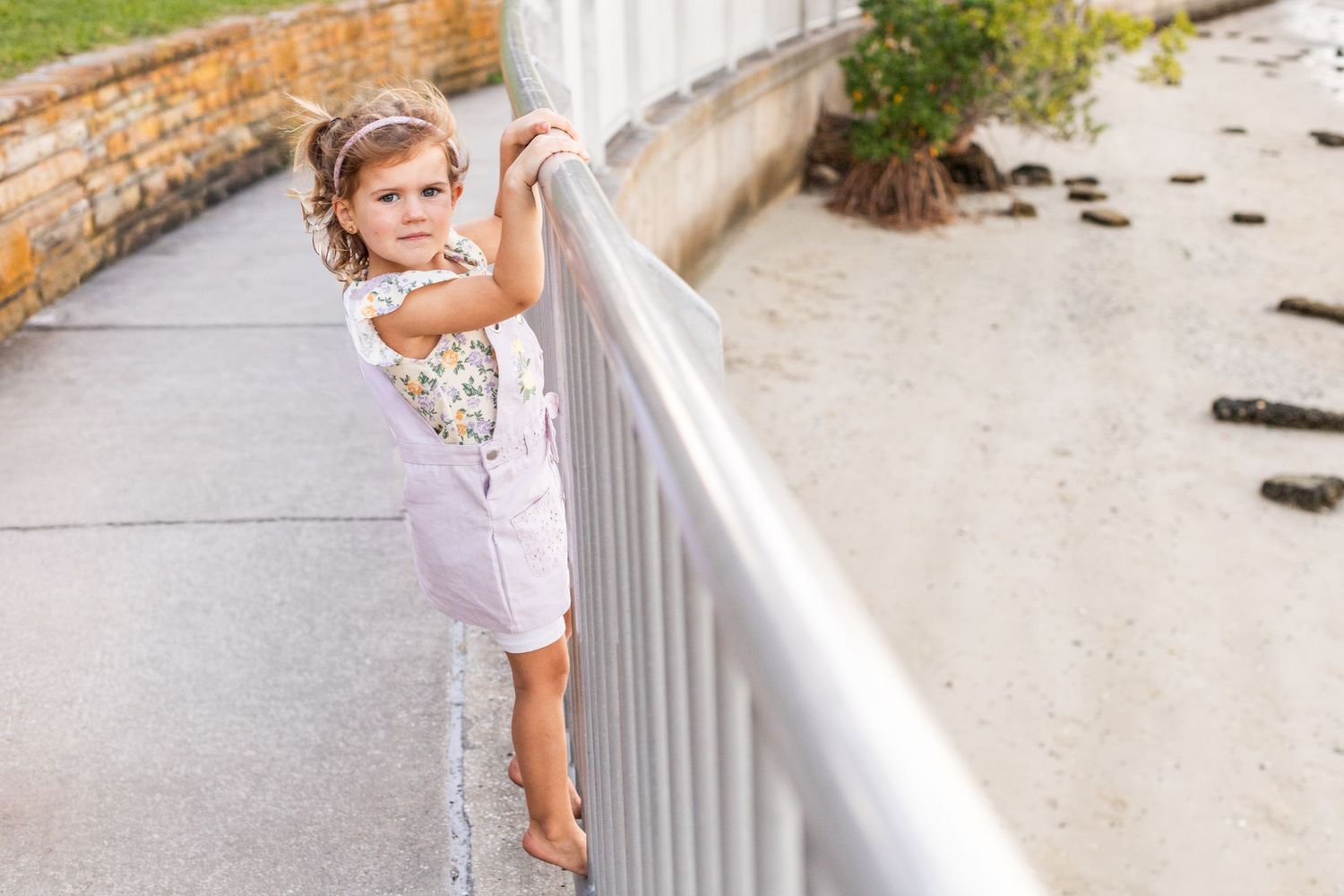 A young toddler girl in pink shorts holds a silver railing on a beachside walkway, looking down at the camera.