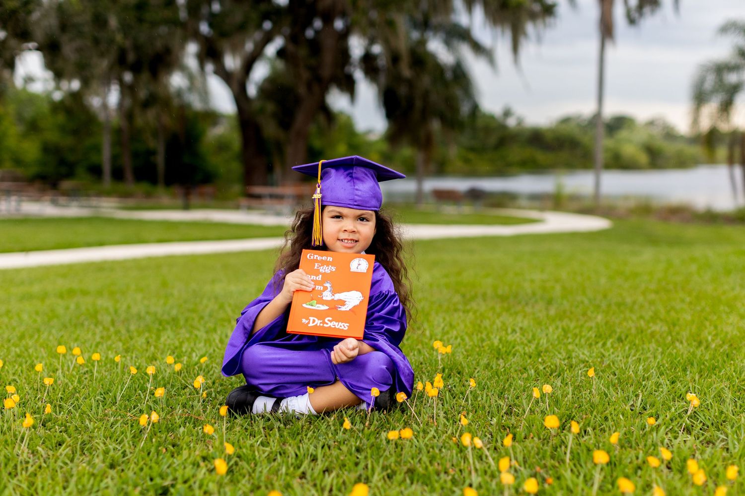 Young graduate in purple cap and gown sitting on grass with yellow flowers holding a book.