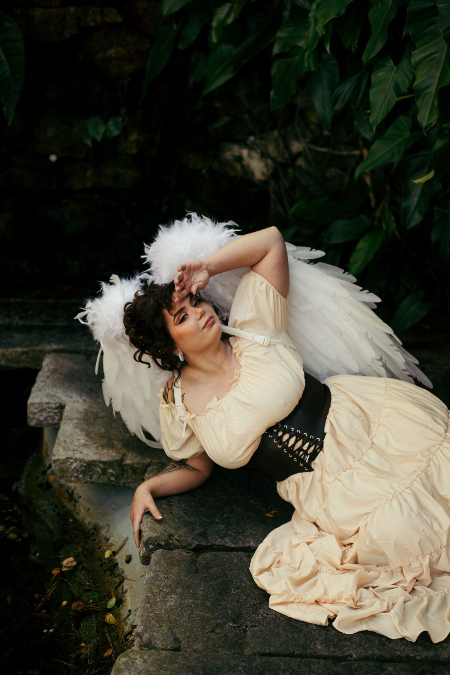 Dramatic garden photoshoot featuring cream dress and white feather wings on stone steps.
