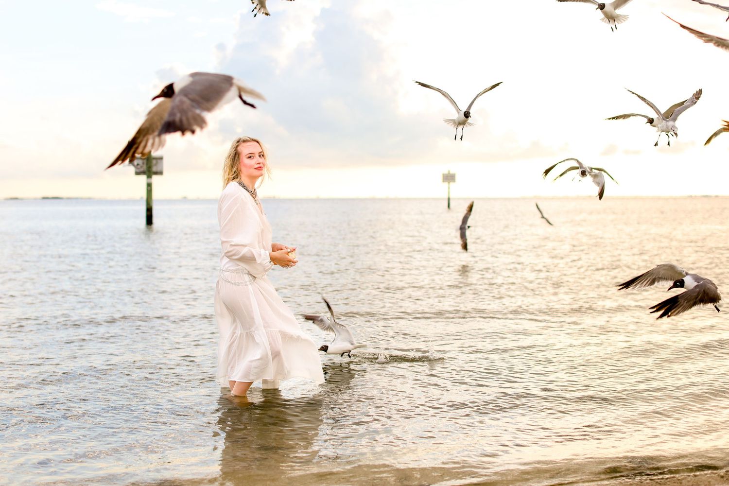Joyful moment feeding seagulls in white dress at the ocean's edge during sunset.