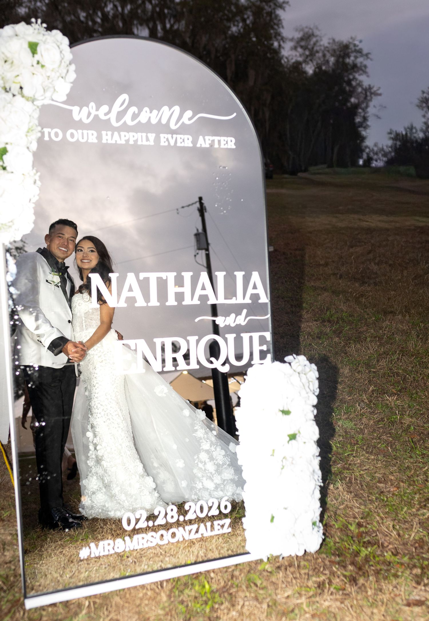 A decorative wedding welcome sign with floral accents displays 'Nathalia and Enrique' with a date.