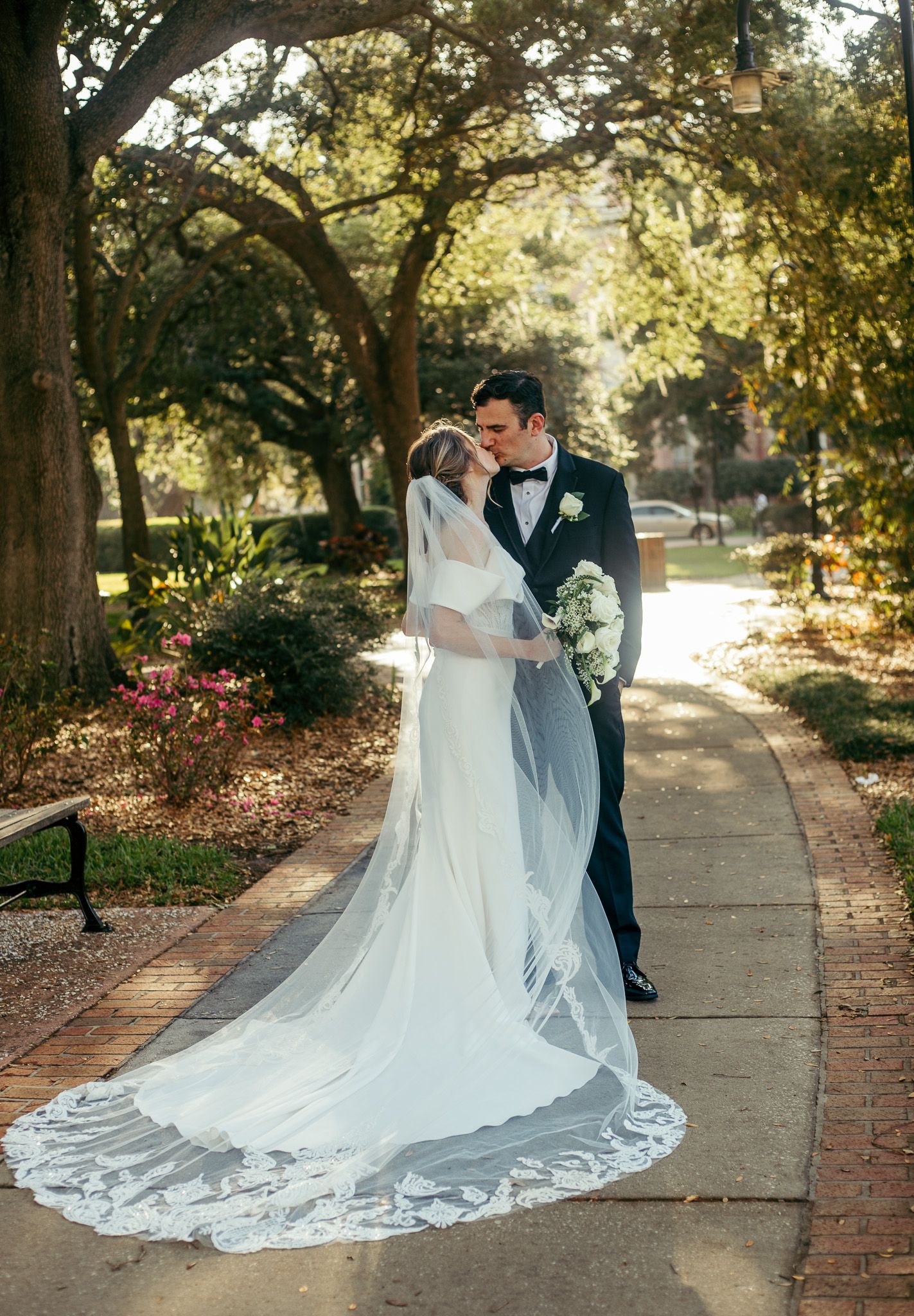 Elegant wedding couple poses on a tree-lined brick pathway with flowing white dress train.