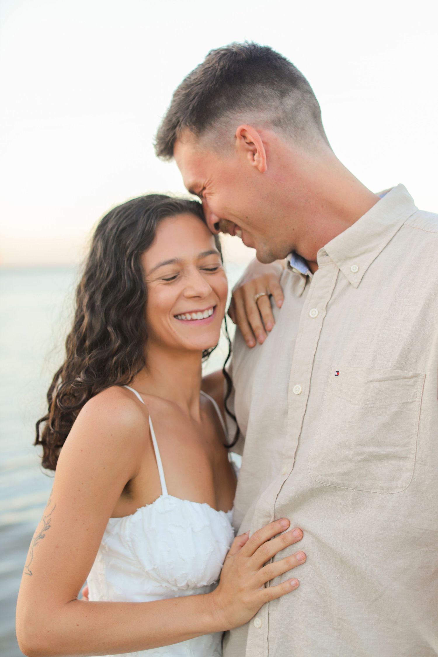 A smiling couple embraces near the water at sunset, woman in white dress with curly hair, man in beige shirt.
