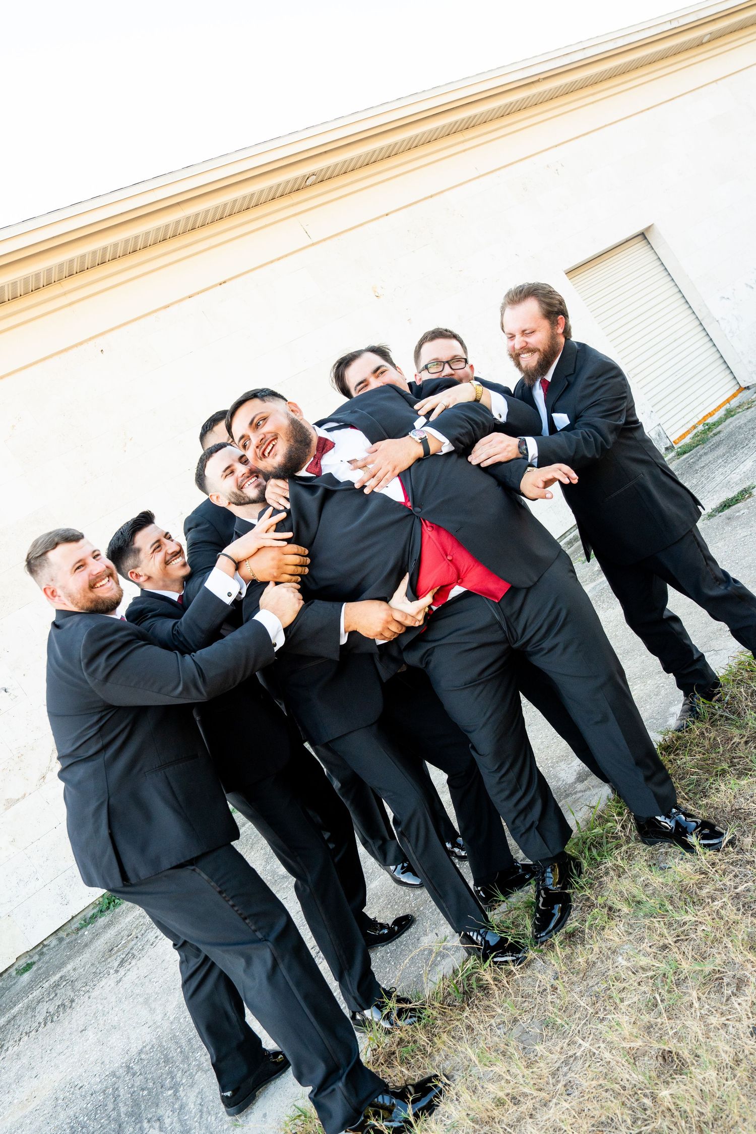 Groomsmen in black suits playfully lifting the groom in a red tie outdoors near a building wall.
