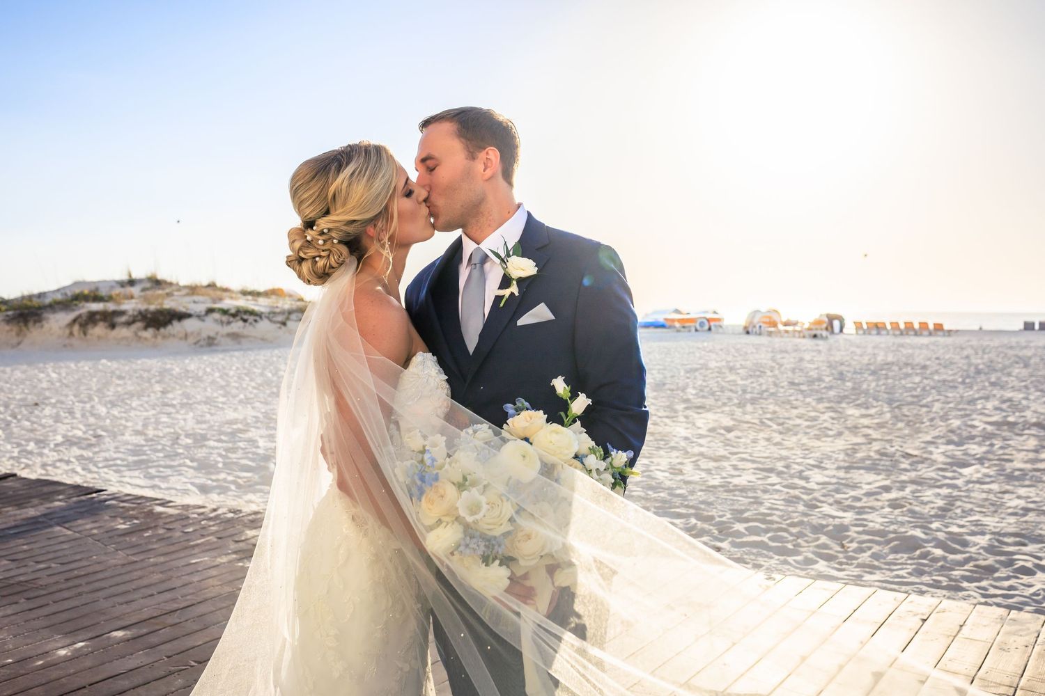 Bride and groom sharing a romantic kiss on a beach boardwalk, bride holding white bouquet with veil flowing in the breeze.