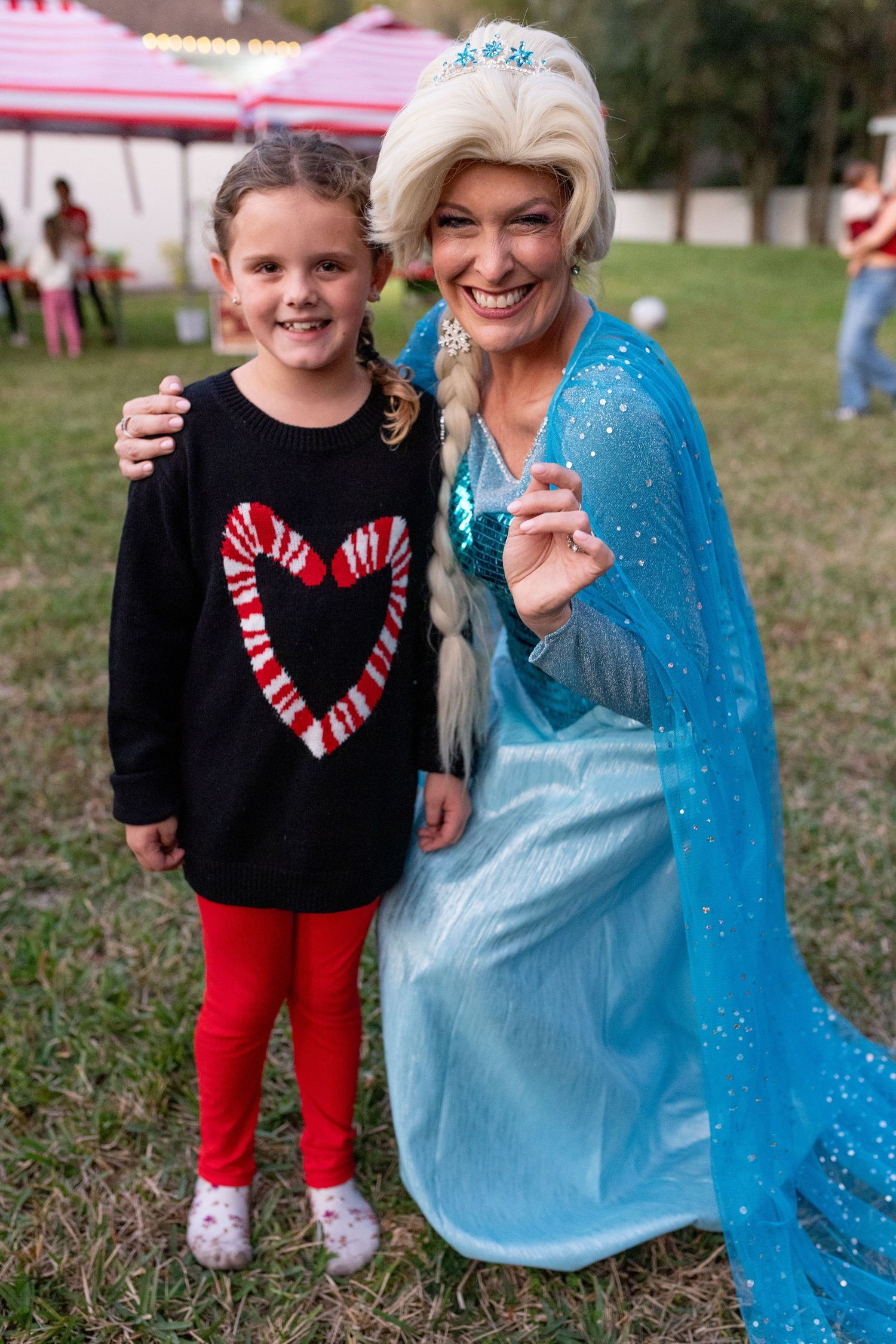 A child in a black shirt with a candy cane heart design poses with someone in a sparkly blue dress outdoors.