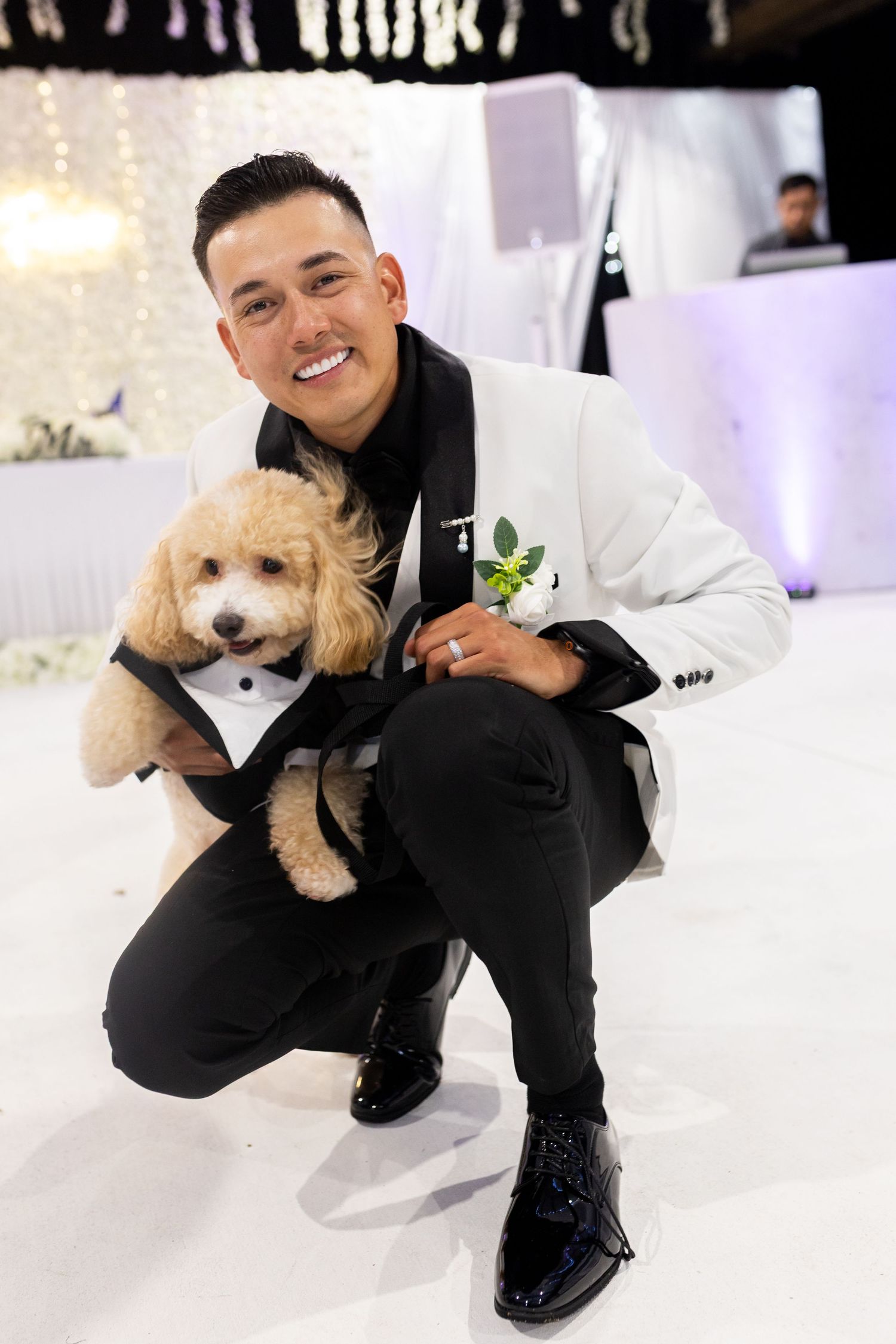 A small dog wearing a tuxedo poses with someone in a white jacket at a formal event.