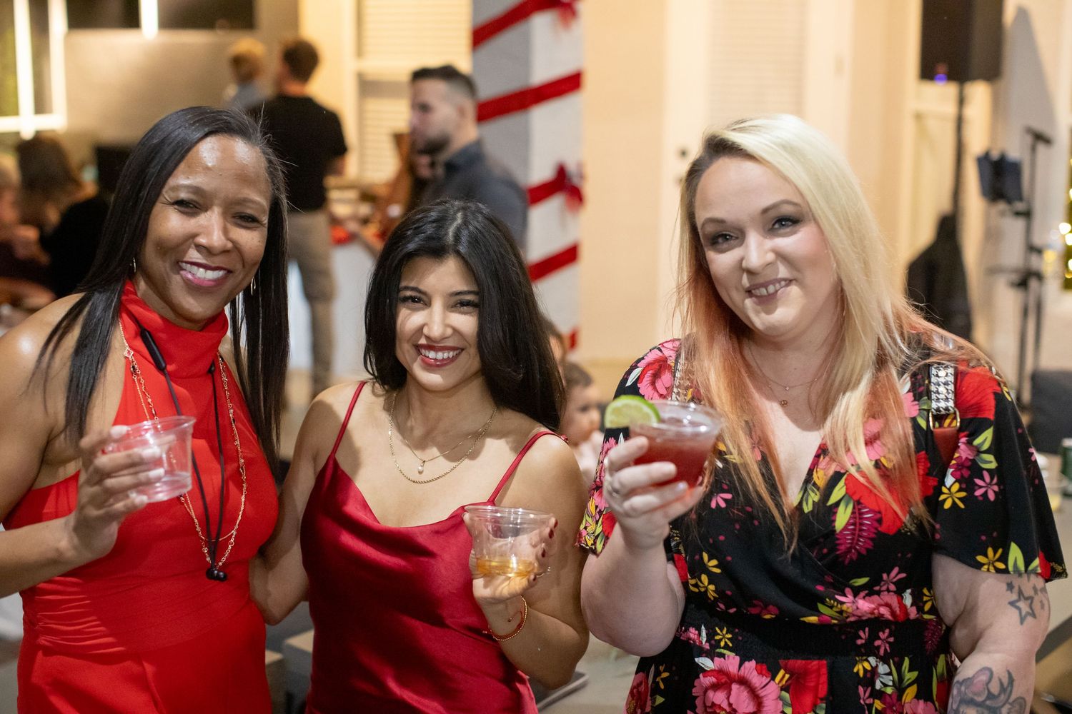Three women in festive attire holding cocktails at holiday party.