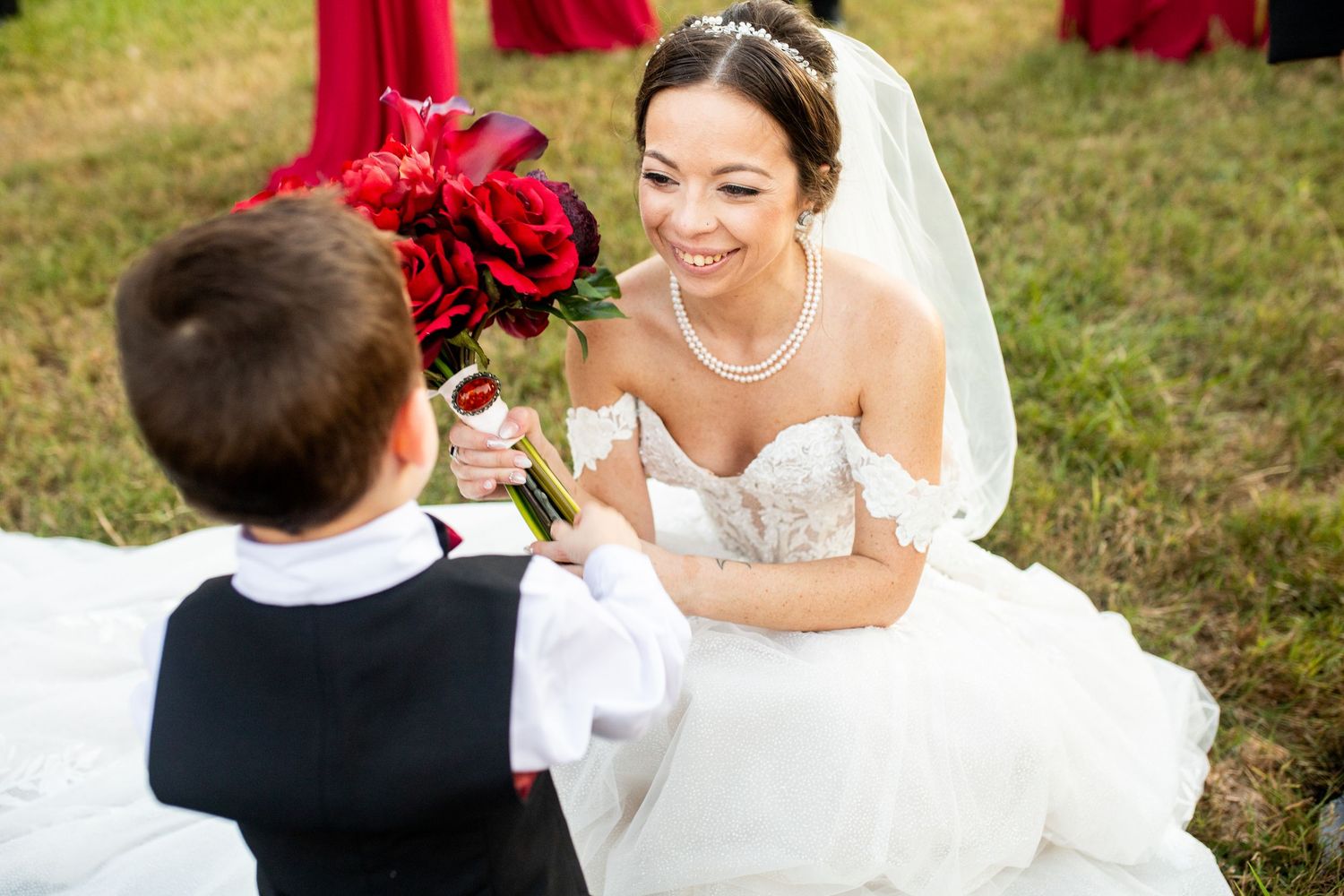 Smiling bride in white gown receives red peony bouquet from young ring bearer at outdoor wedding ceremony.