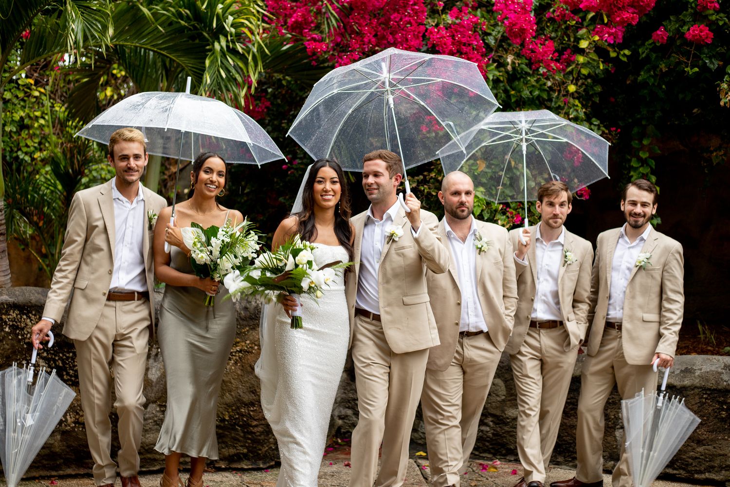 Wedding party with clear umbrellas in beige suits, bride holding white bouquet, posing outdoors with tropical flowers.