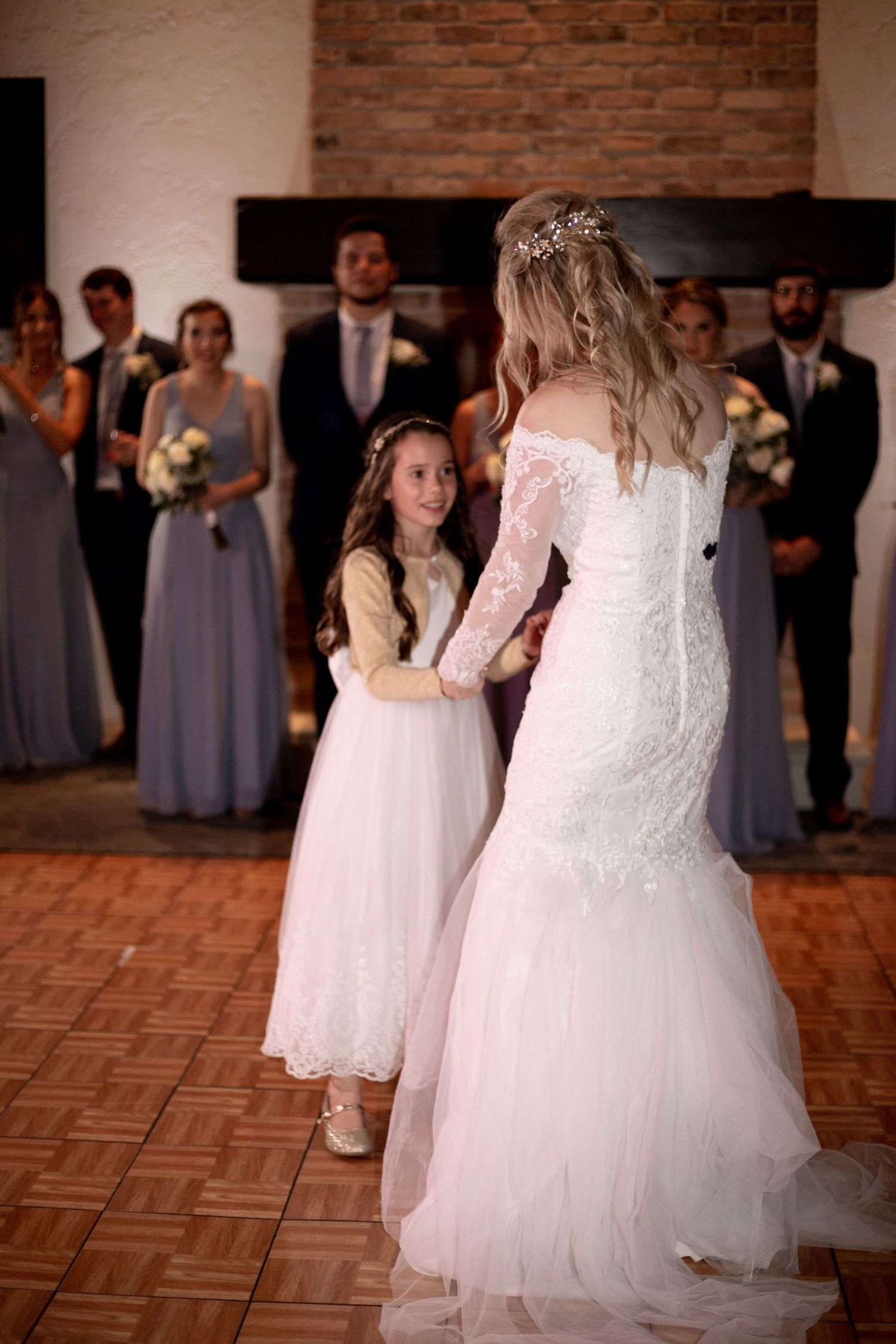 Bride in lace gown dancing with flower girl in white dress at wedding reception with bridal party watching.