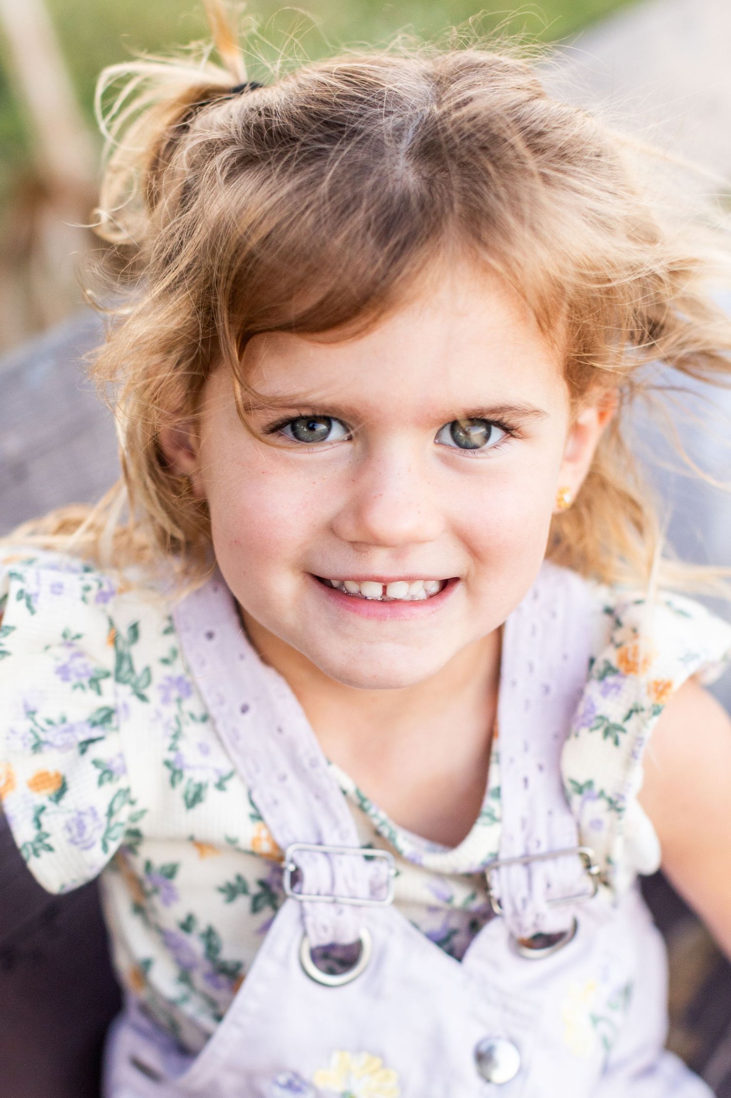 Smiling young girl with curly blonde hair and green eyes wearing floral top and lavender overalls outdoors.