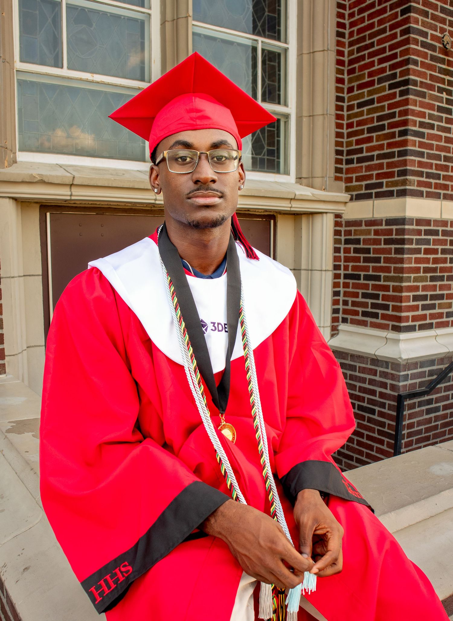Graduate in red academic regalia sits thoughtfully outside brick university building.
