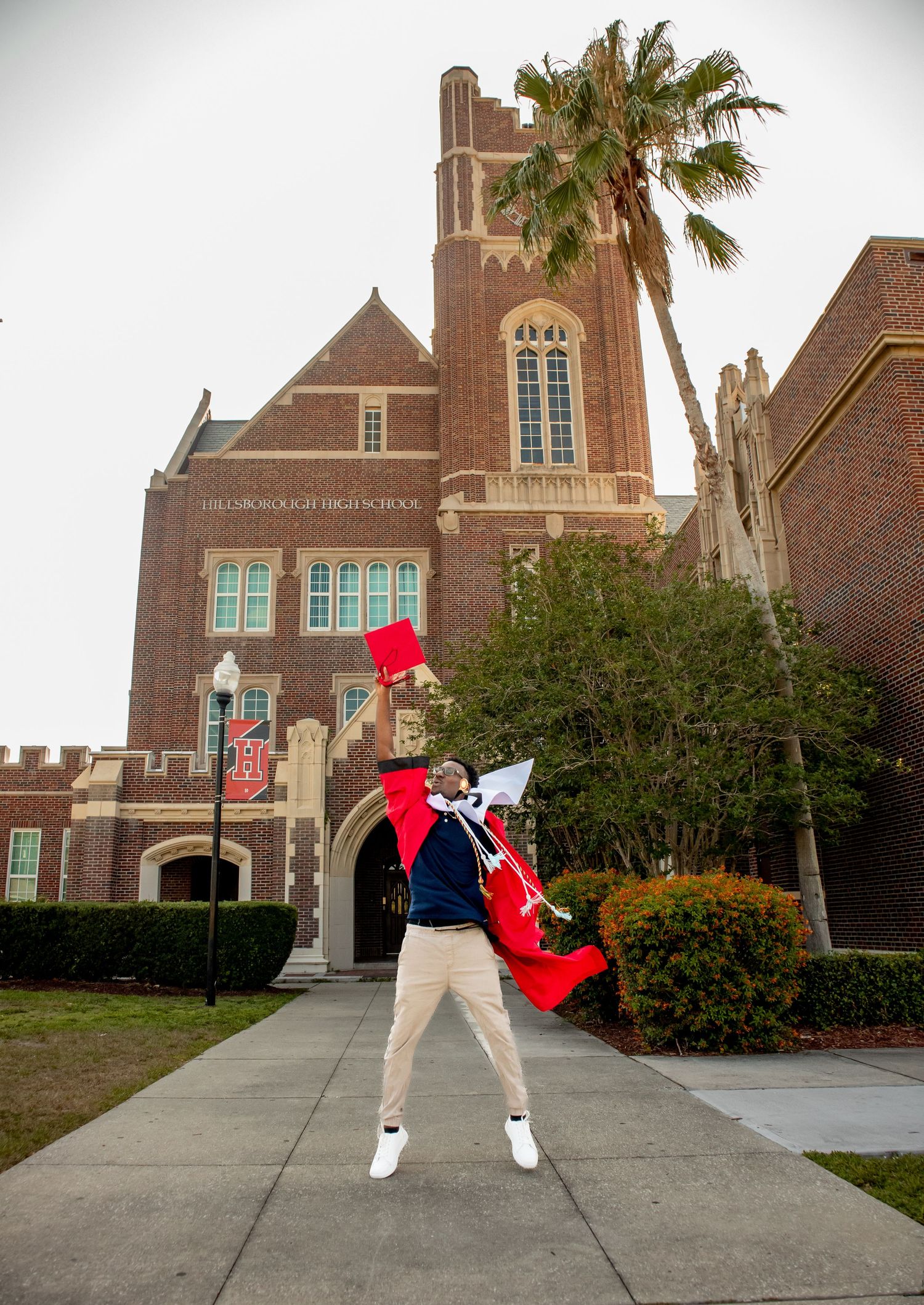 Graduates perform energetic dance moves in red robes outside brick building.