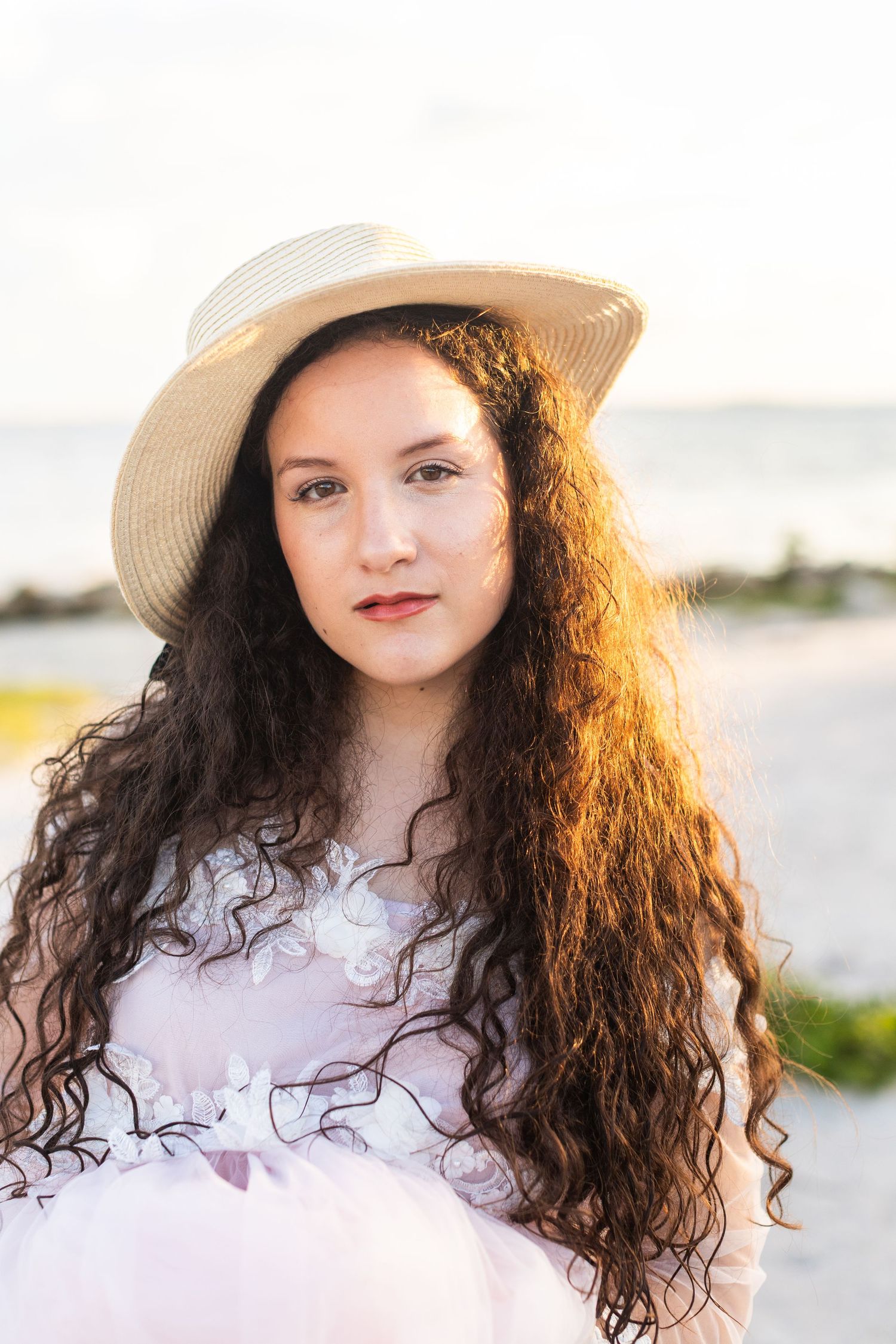 Summer beach portrait with straw hat, white lace dress and natural curls in golden hour light.