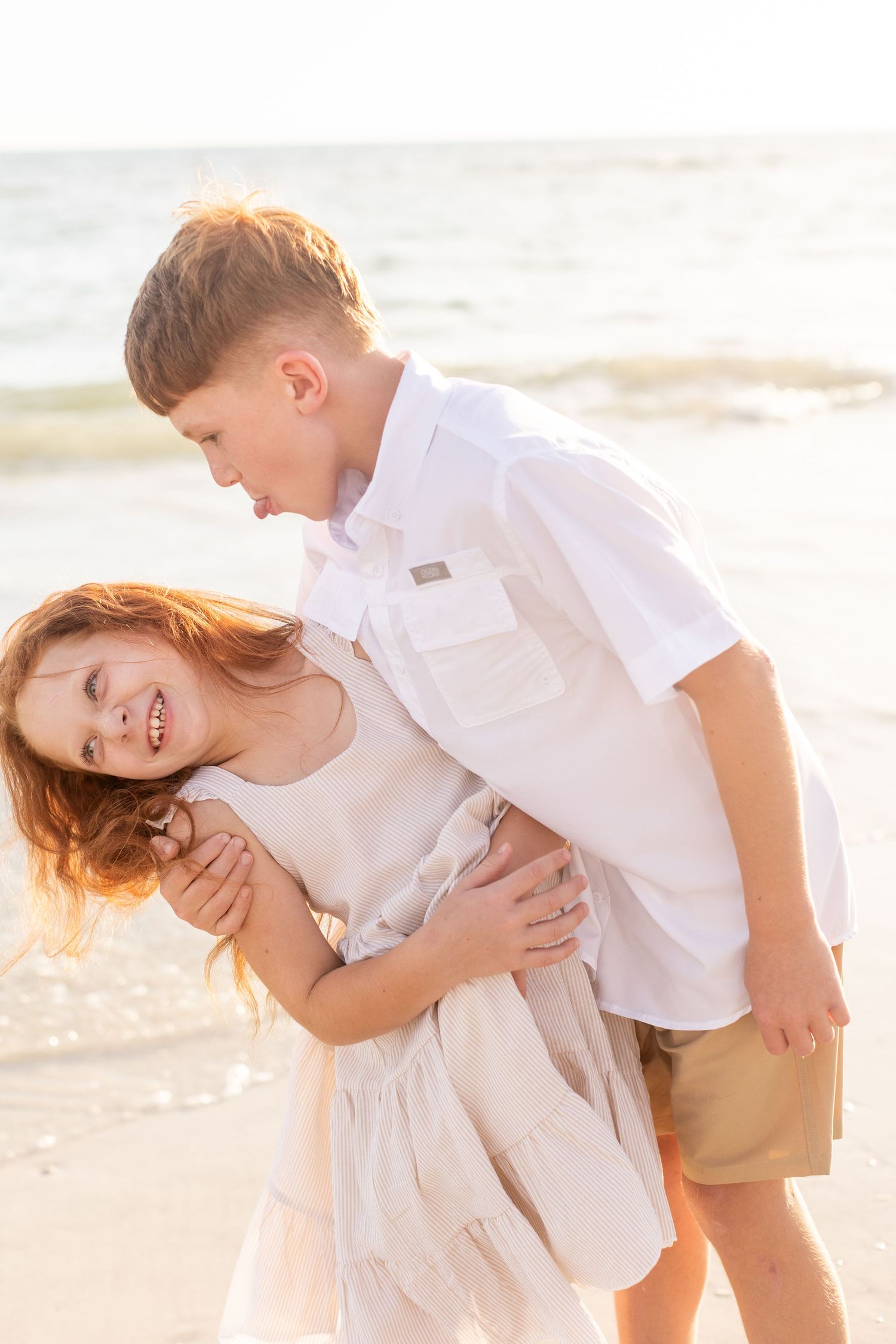 Playful beach photography of siblings in light colored clothing at golden hour.