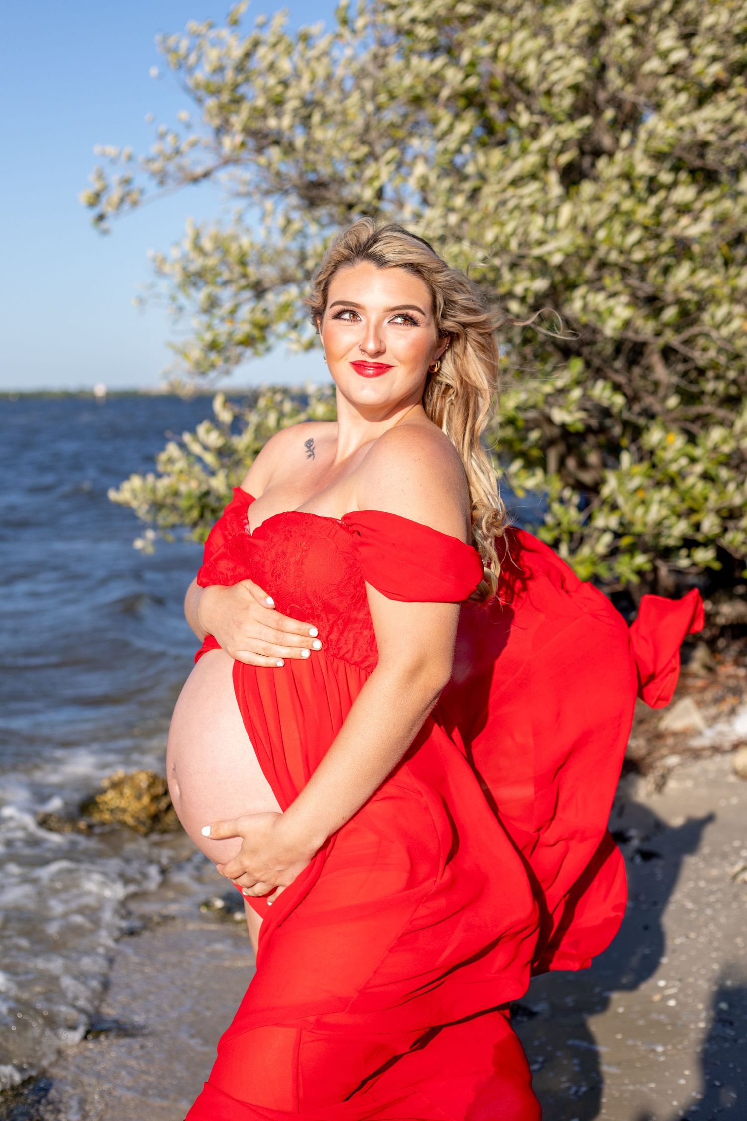 A pregnant woman in a flowing red dress poses by the ocean.