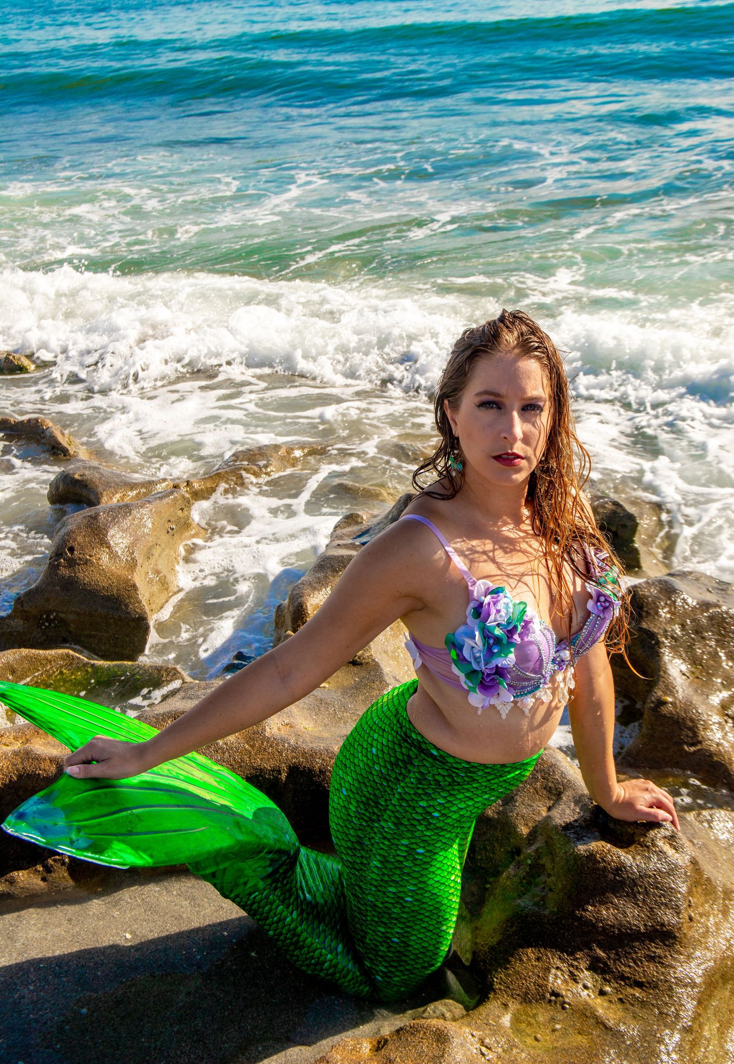 A mermaid costume photoshoot on rocky coastal rocks with bright turquoise ocean waves in the background.