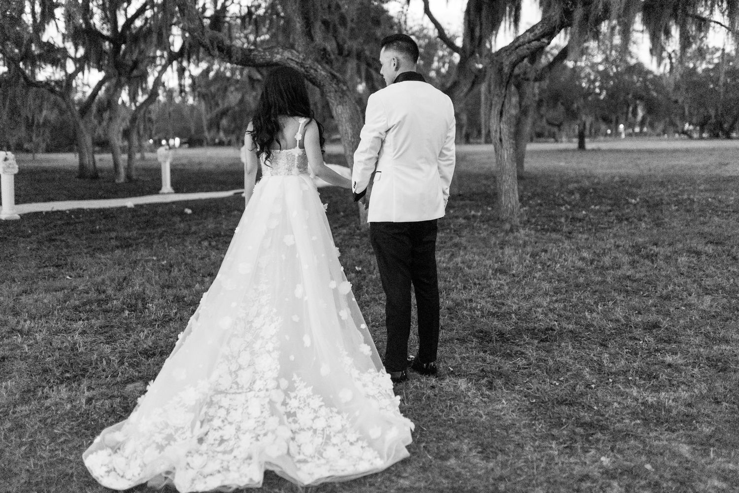 A couple in wedding attire walks together on grass beneath Spanish moss-draped trees.
