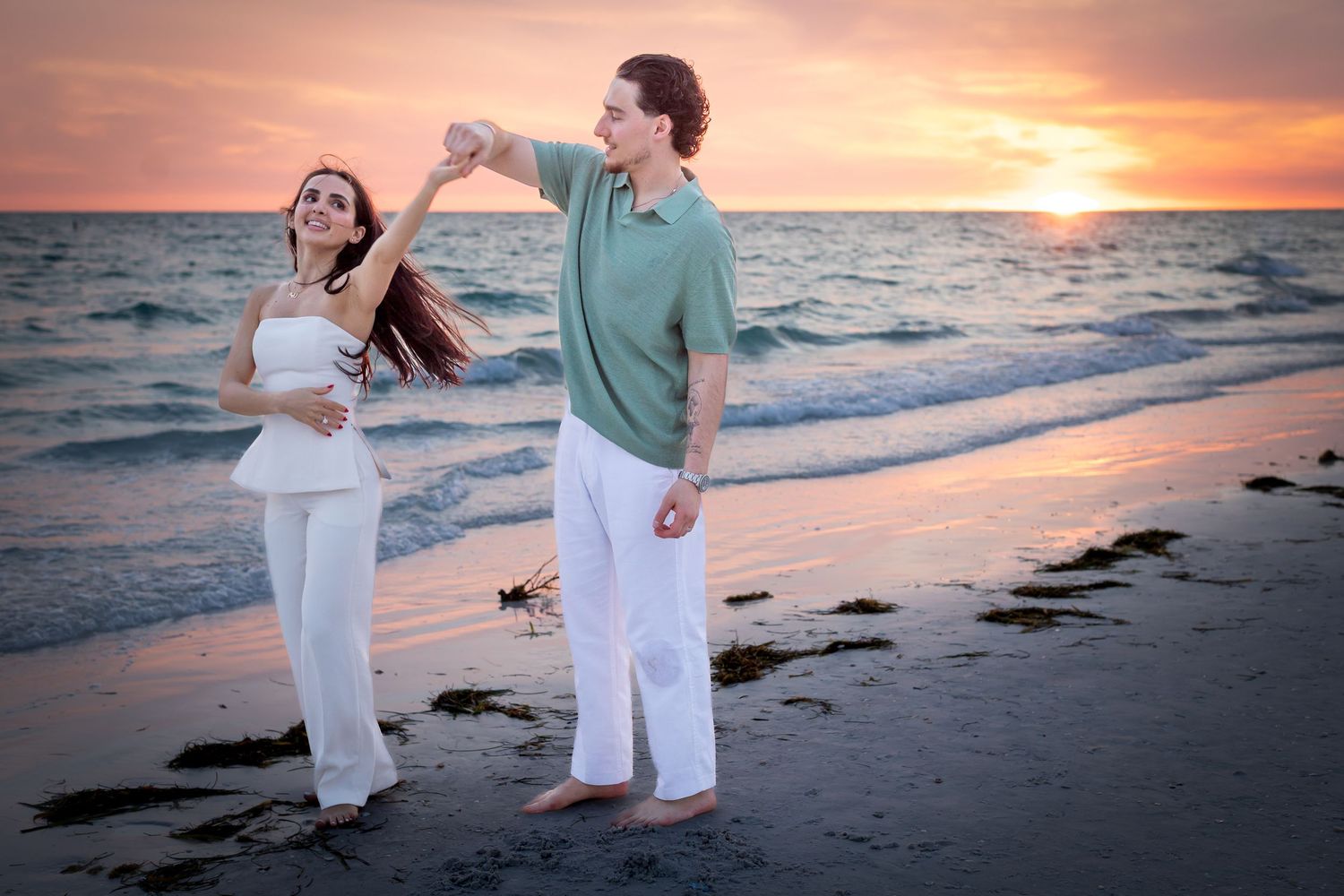 Couple dancing romantically on a beach at sunset, woman in white outfit spinning gracefully by the ocean waves.