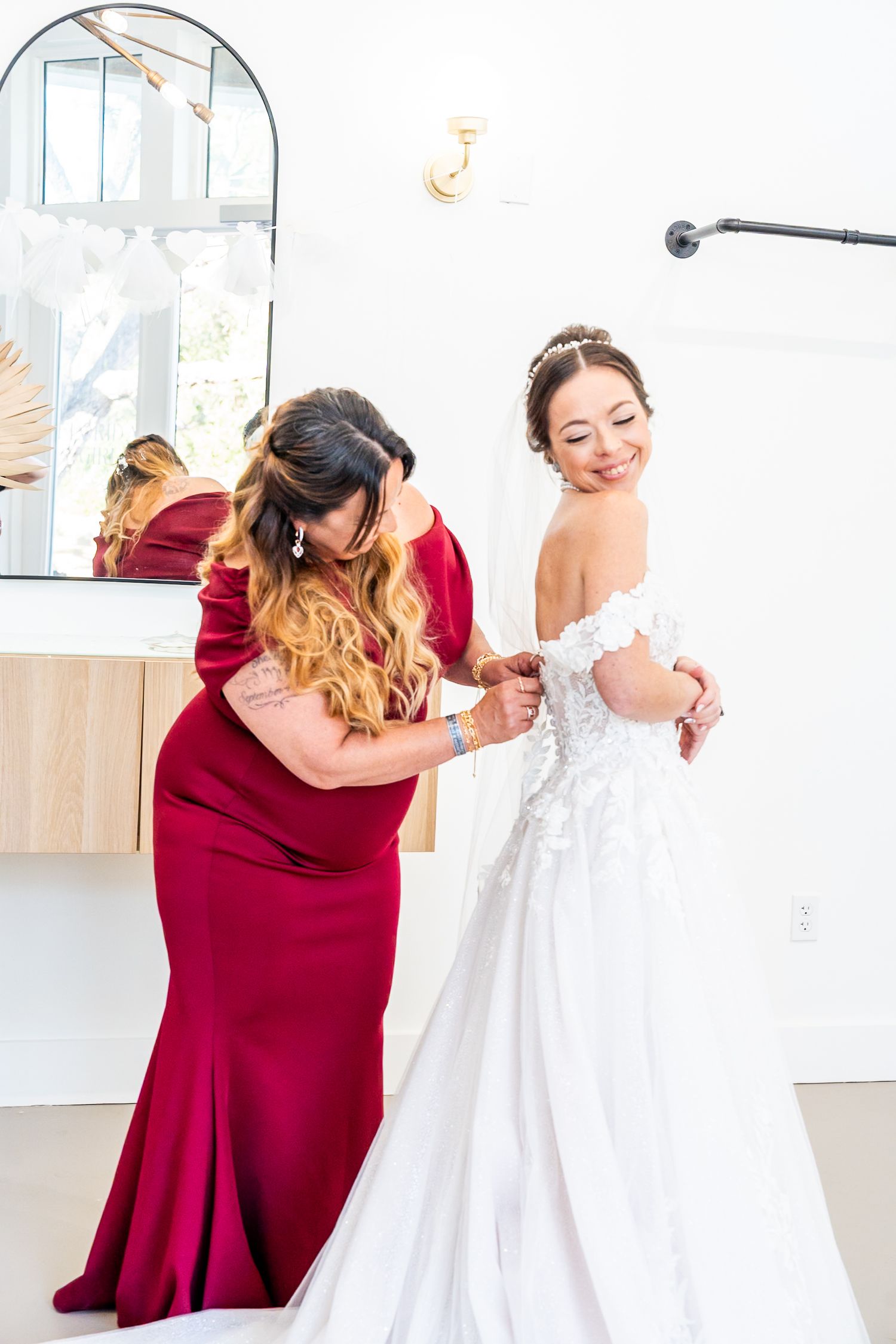 A woman in a red gown helps a smiling bride button her white lace wedding dress in a bright bridal suite.