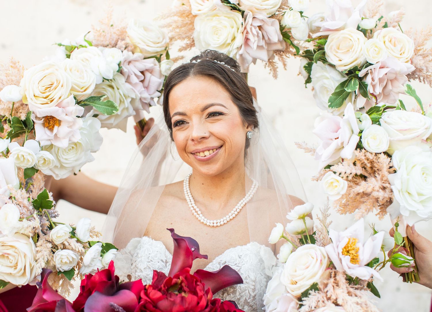 Smiling bride in veil and pearl necklace framed by white and blush floral arch holding red bouquet.