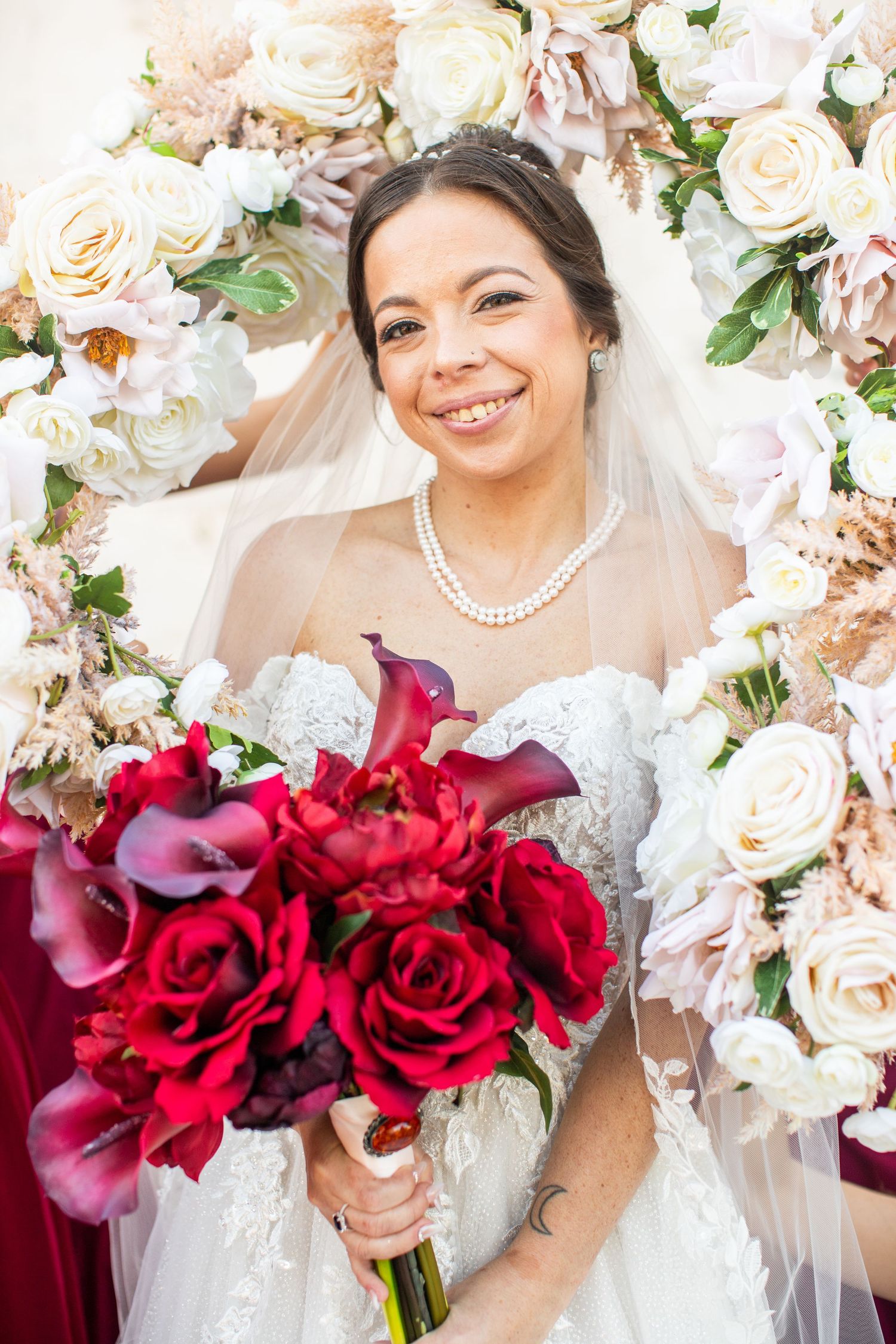 Smiling bride in white lace gown holds red rose bouquet, wearing pearl necklace and veil before floral arch.