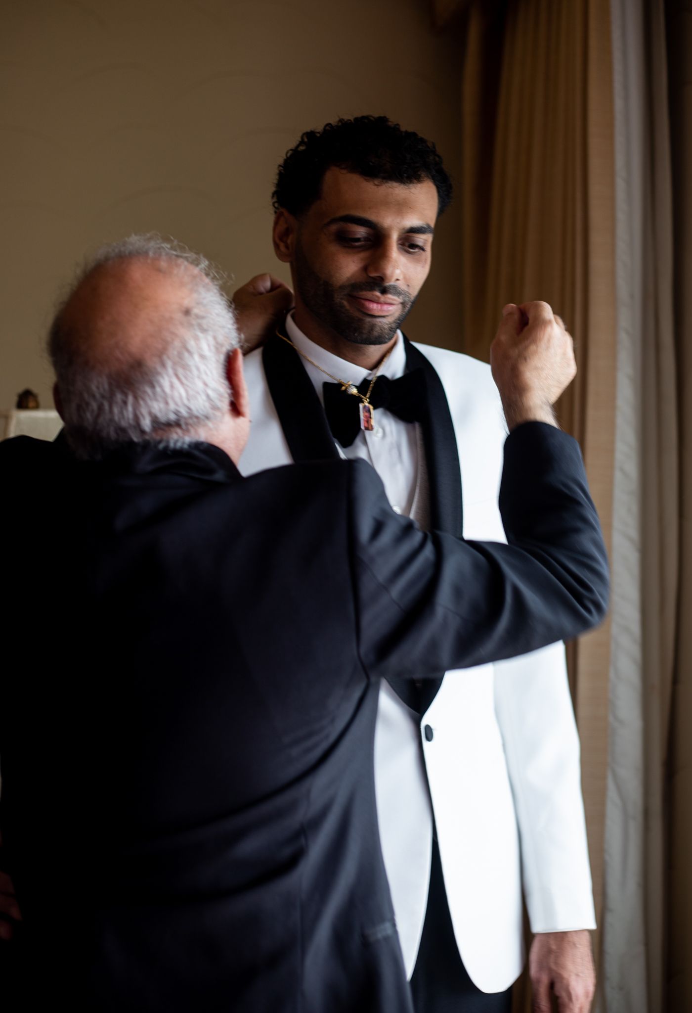 Older man helping a young groom in a white tuxedo jacket adjust his bow tie before a wedding ceremony.
