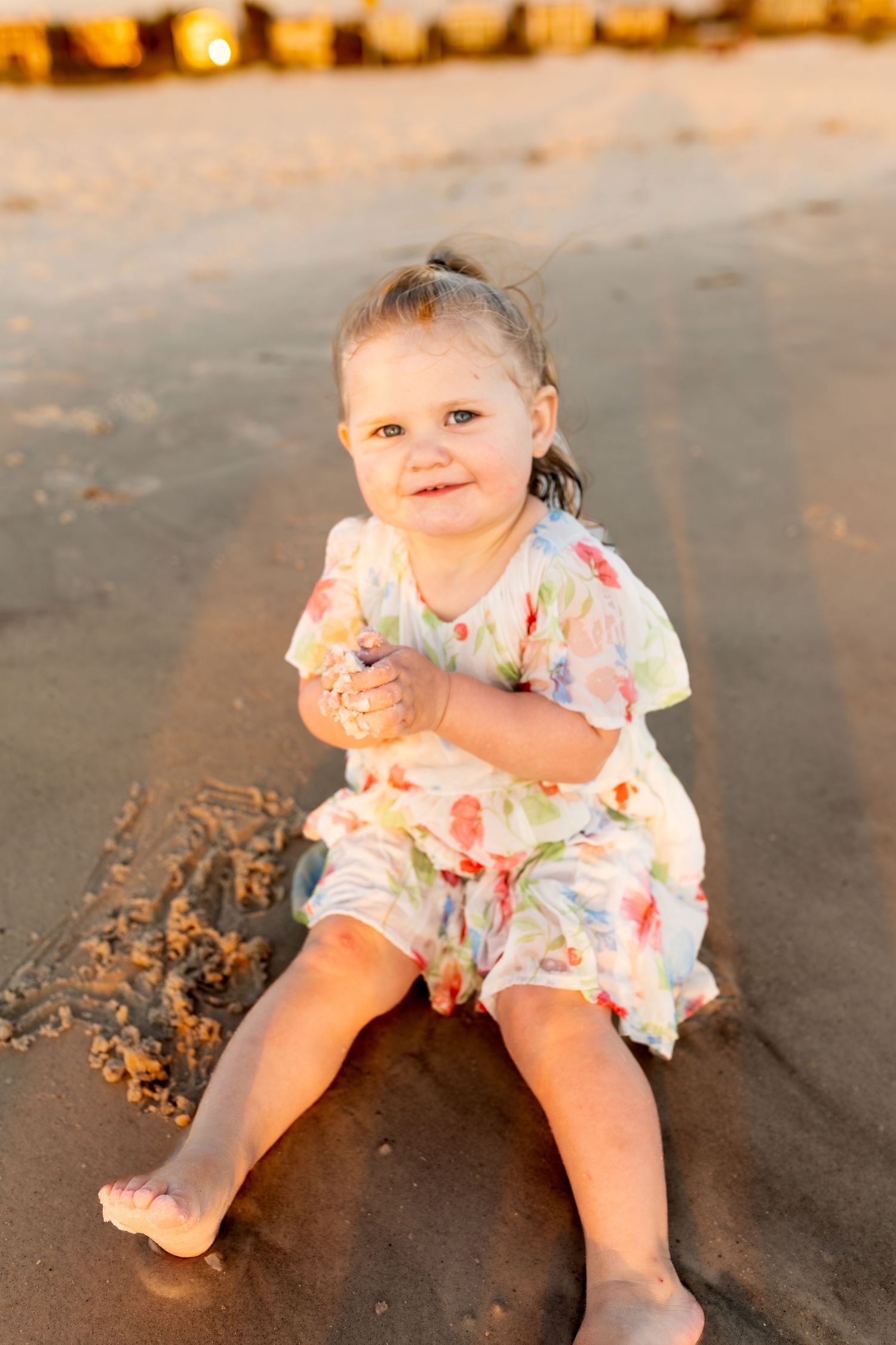 Cute toddler girl in floral dress sitting on sandy beach at golden hour, smiling and playing with sand.