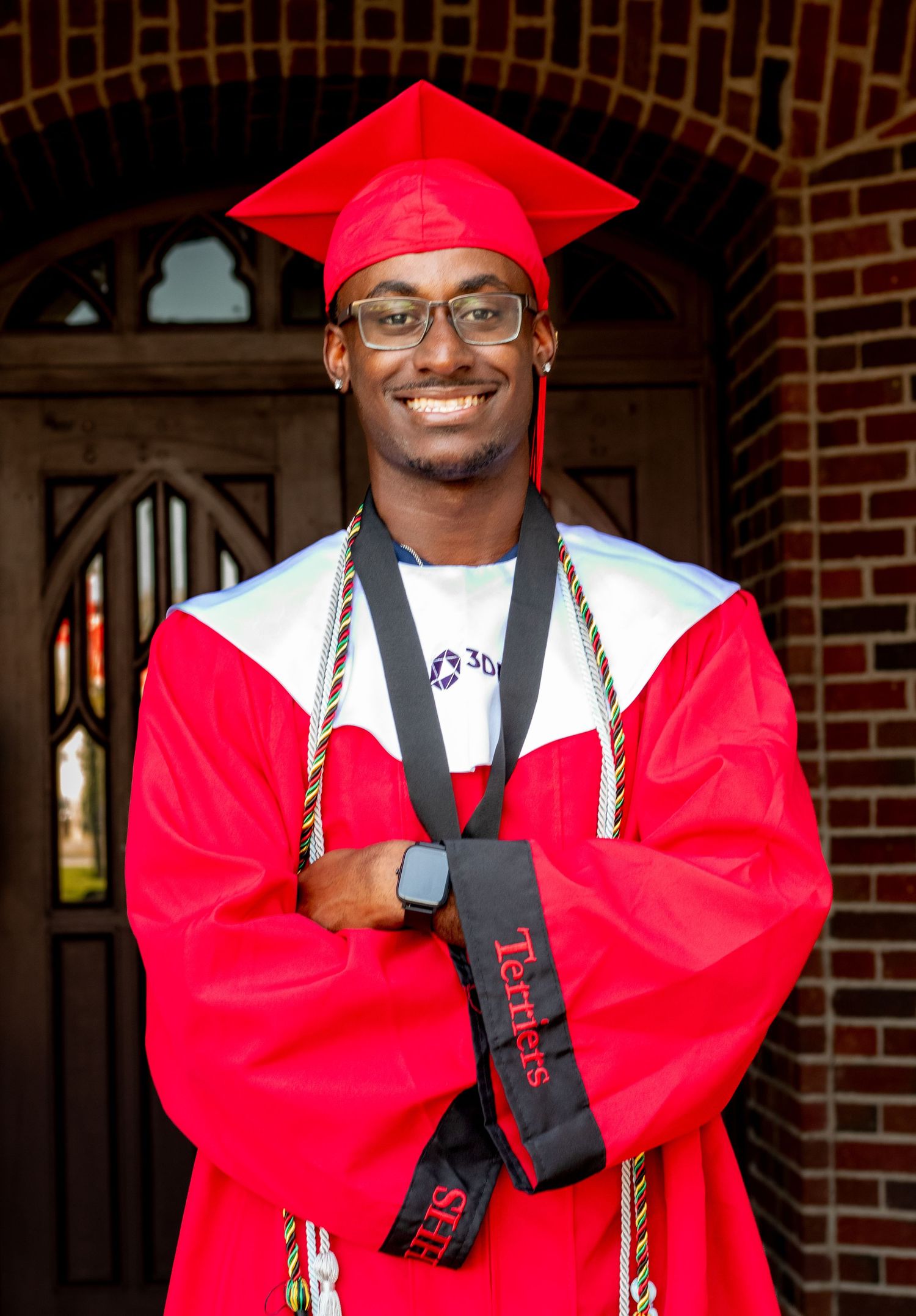 Graduate wearing red and white regalia smiles in front of arched brick doorway.