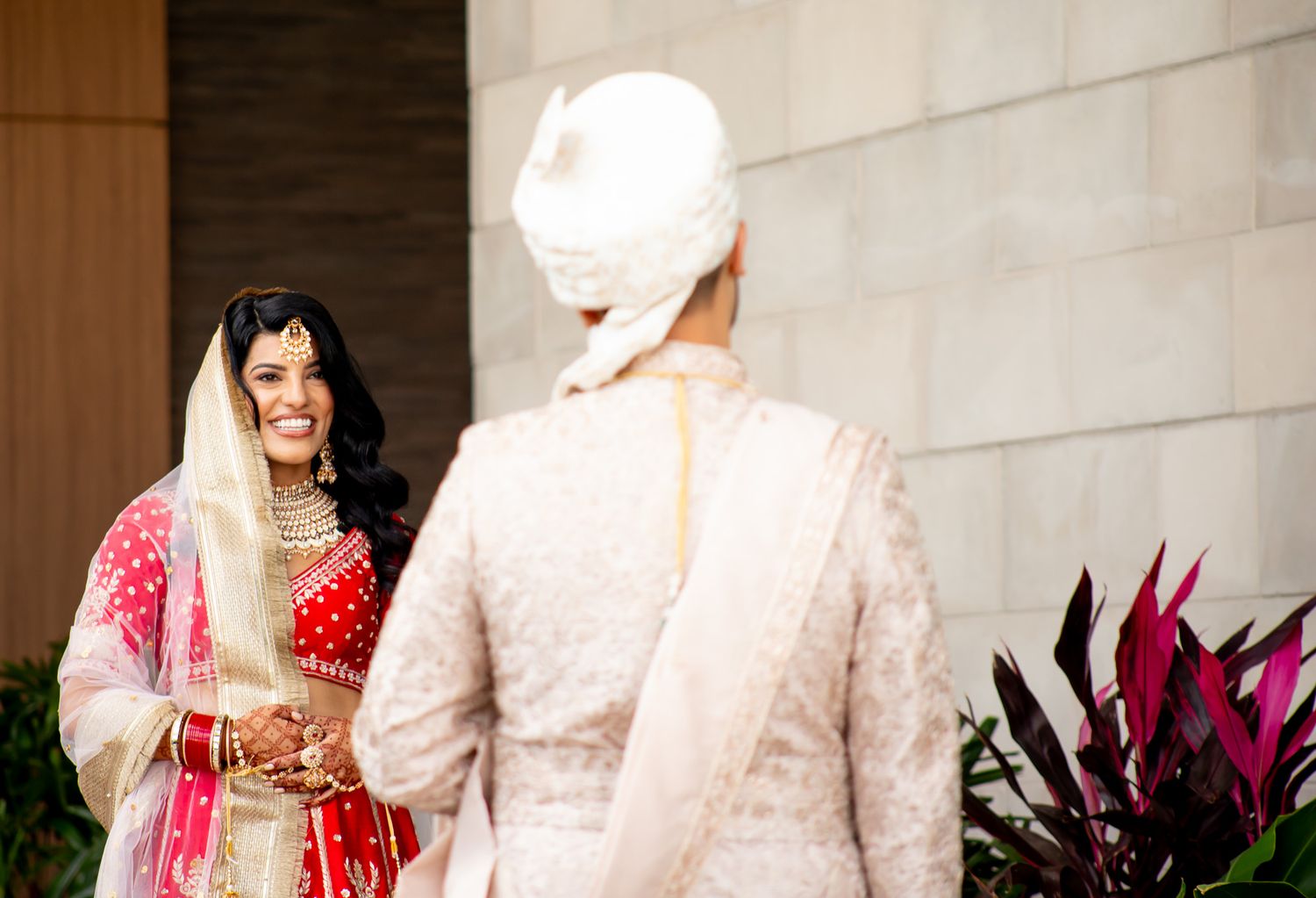 Traditional Indian wedding couple in elegant red and gold attire pose together against a light wall.