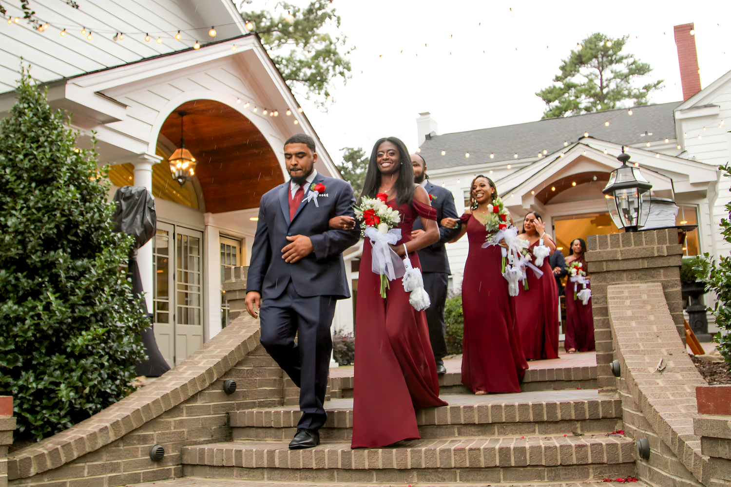 Wedding party in burgundy dresses poses on brick steps outside a white colonial-style building.