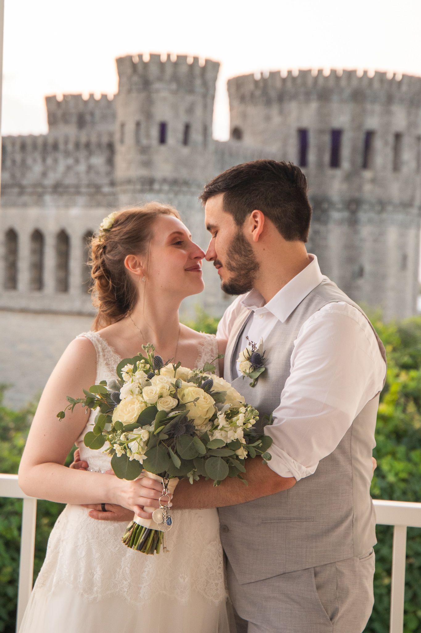Wedding couple embraces on balcony with castle-like architecture in background.