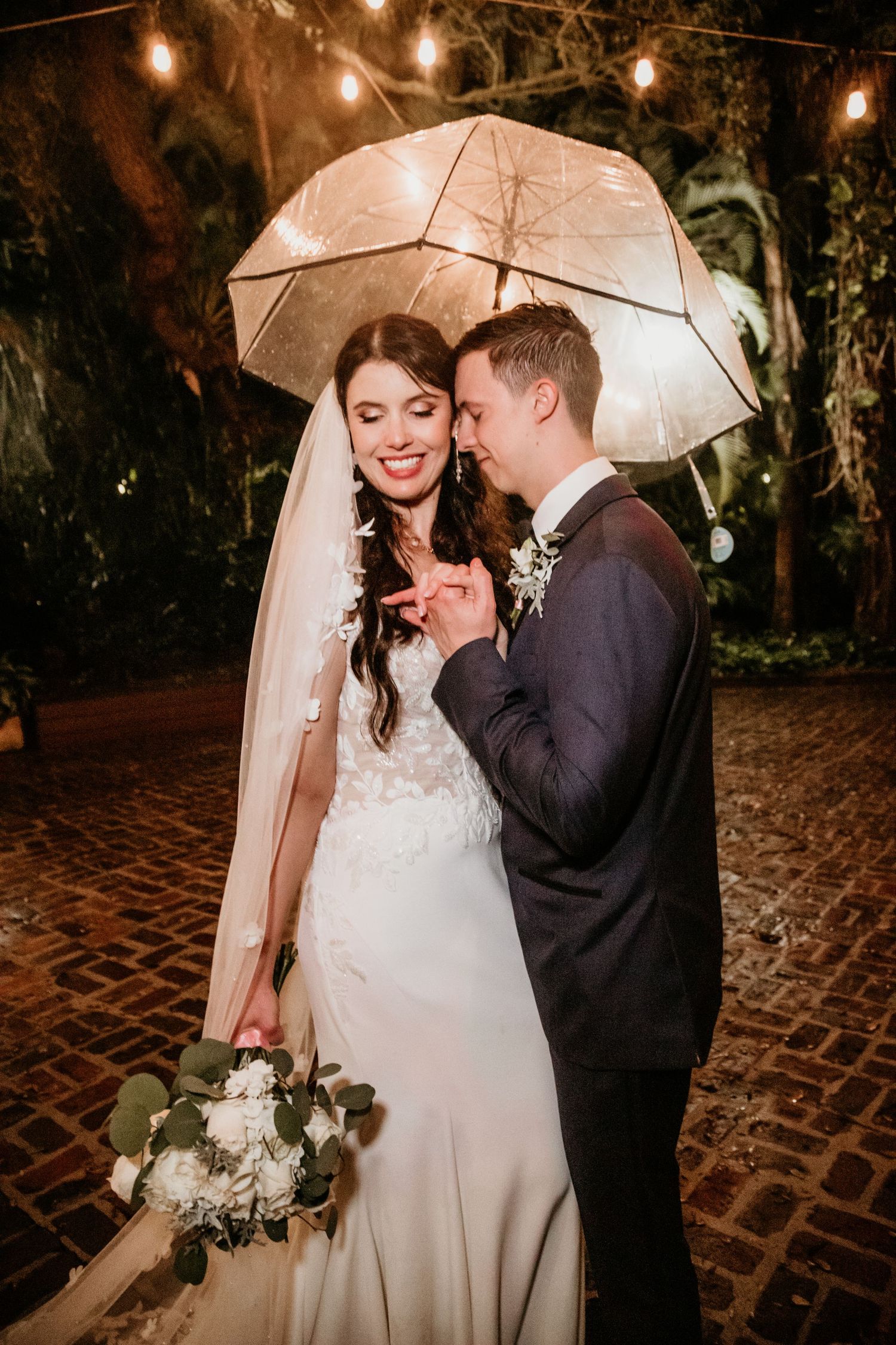 Intimate wedding couple moment under clear umbrella with string lights glowing overhead on brick patio.