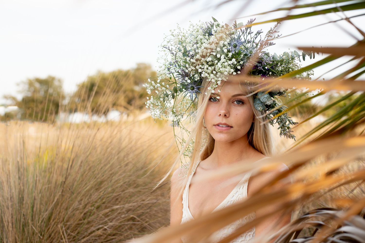 Dreamy portrait through palm fronds featuring delicate floral crown and white dress.