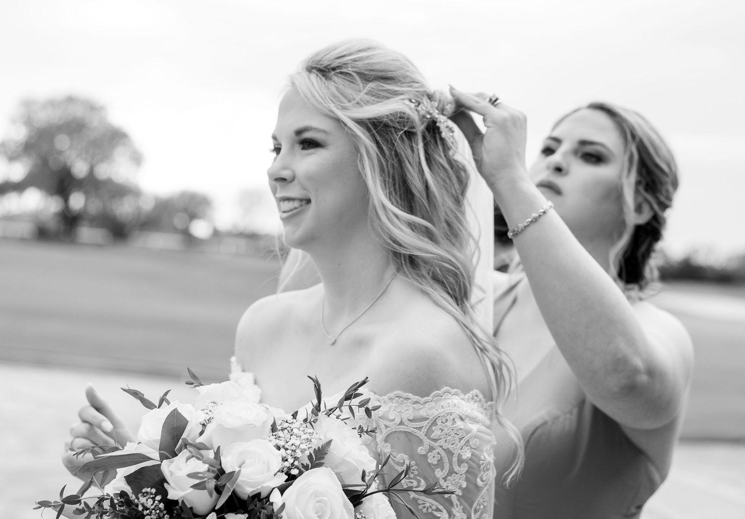 Black and white photo of a bride holding a bouquet while a bridesmaid adjusts her hair outdoors.