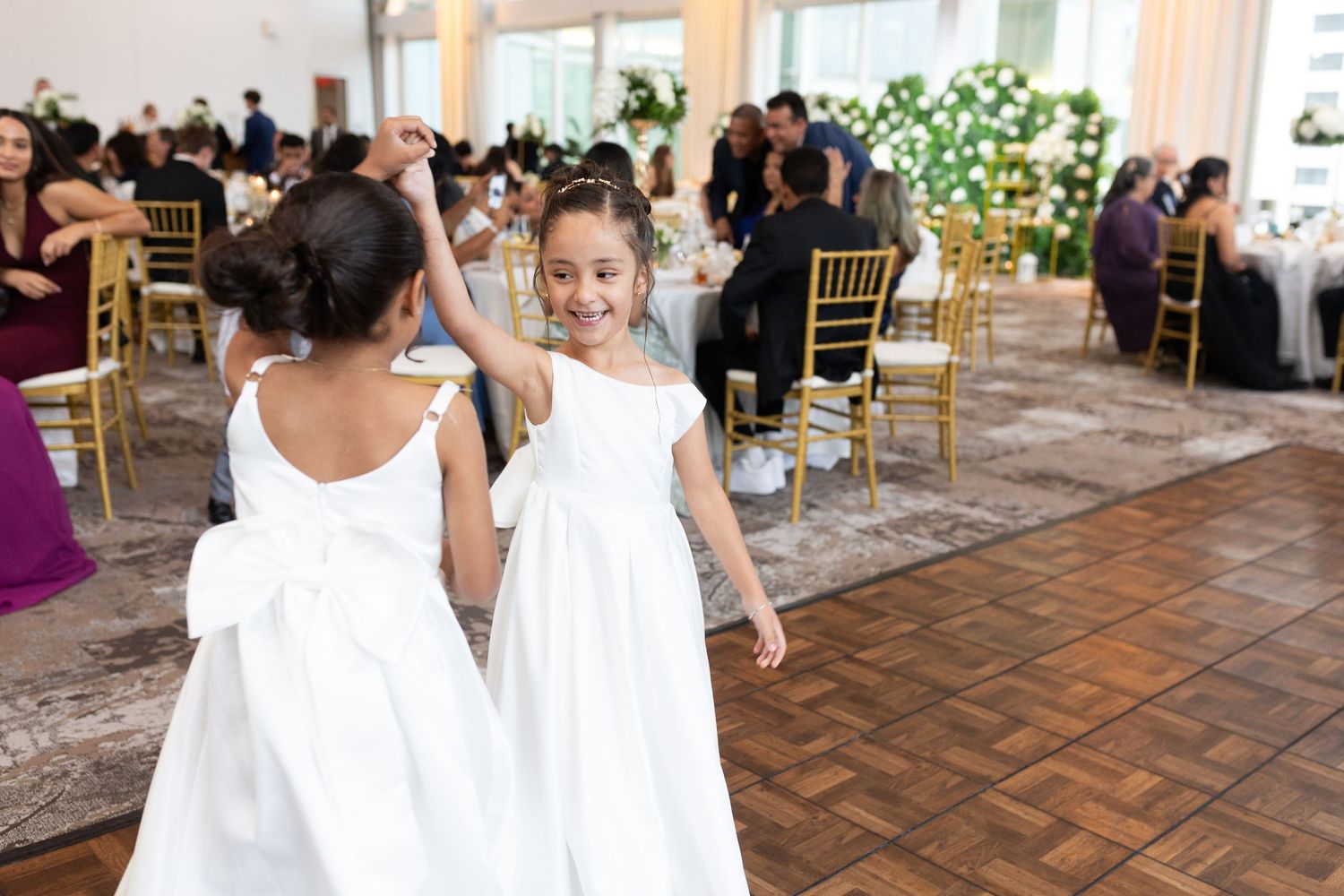 Two flower girls in white dresses with bows dance joyfully at an elegant wedding reception with gold chairs.
